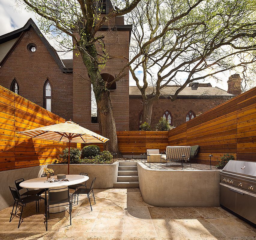 This sophisticated multi-level patio features a dining area with a striped umbrella and a raised lounge space, all enclosed by warm-toned horizontal wood fencing. The design incorporates modern concrete retaining walls and stone pavers, creating a seamless transition between the dining and grilling zones. A large, mature tree provides a natural canopy, framing the view of the historic brick church in the background for a unique urban oasis feel.