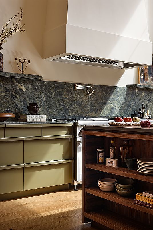 This sophisticated kitchen features a striking dark green marble backsplash paired with muted olive-toned cabinetry and warm wood shelving. A large, minimalist range hood anchors the cooking area, while the foreground showcases an open-shelf wooden island stocked with ceramic bowls and kitchen essentials. The scene is bathed in soft, natural light, creating an inviting and high-end culinary atmosphere.