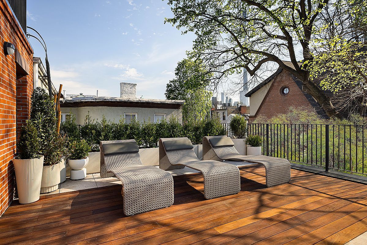 This inviting rooftop deck features warm-toned wooden decking and three modern, woven lounge chairs arranged for relaxation. The space is framed by lush green hedges and potted plants, with a black metal railing overlooking a scenic urban backdrop. The perspective is a wide, eye-level shot that captures the serene atmosphere and the blend of private outdoor living with city views.
