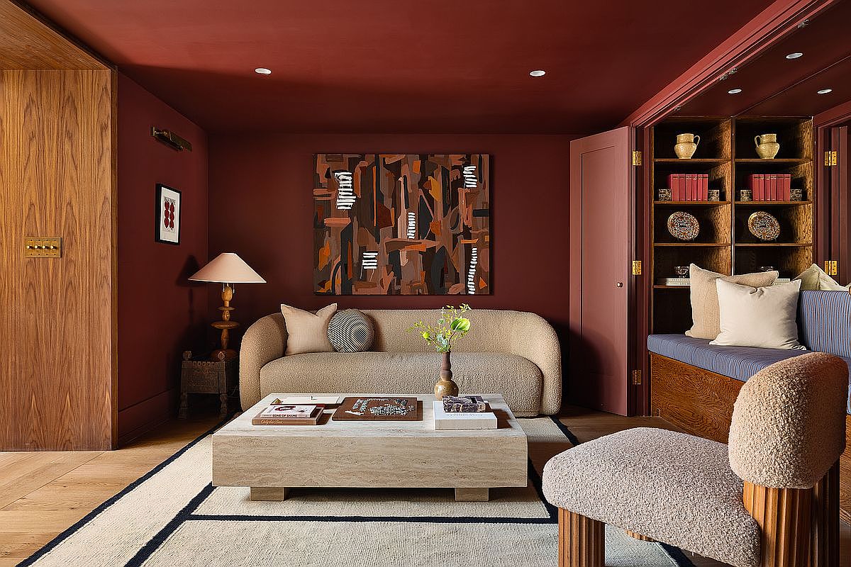 This sophisticated living room features deep burgundy walls and ceiling, creating a cozy and intimate atmosphere. The space is anchored by a textured, cream-colored curved sofa and a large, minimalist travertine coffee table, complemented by a plush area rug and a matching accent chair. Built-in wooden shelving units and warm, ambient lighting from a table lamp add depth and character to this stylish, modern interior.