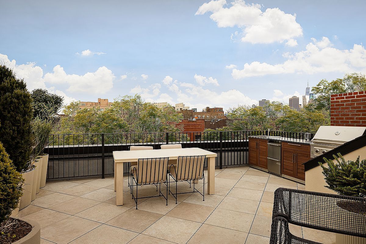 This spacious rooftop terrace features a modern outdoor dining set and a built-in stainless steel kitchen area, perfect for entertaining. The area is paved with large stone tiles and enclosed by a sleek black metal railing, offering panoramic views of the surrounding cityscape and trees. The perspective is a wide-angle shot from the terrace level, capturing the inviting atmosphere of an urban outdoor living space.