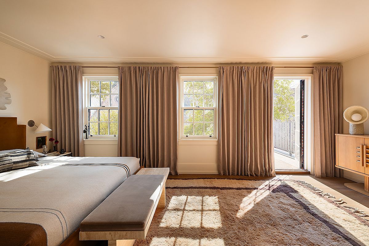 This elegant primary bedroom features a neutral color palette, a plush textured area rug, and a sleek wooden bench at the foot of the bed. Large windows and a glass door leading to a balcony allow for ample natural light, creating a bright and airy atmosphere. The room is styled with sophisticated, minimalist decor, including a modern wooden dresser and subtle wall lighting, offering a serene and luxurious retreat.