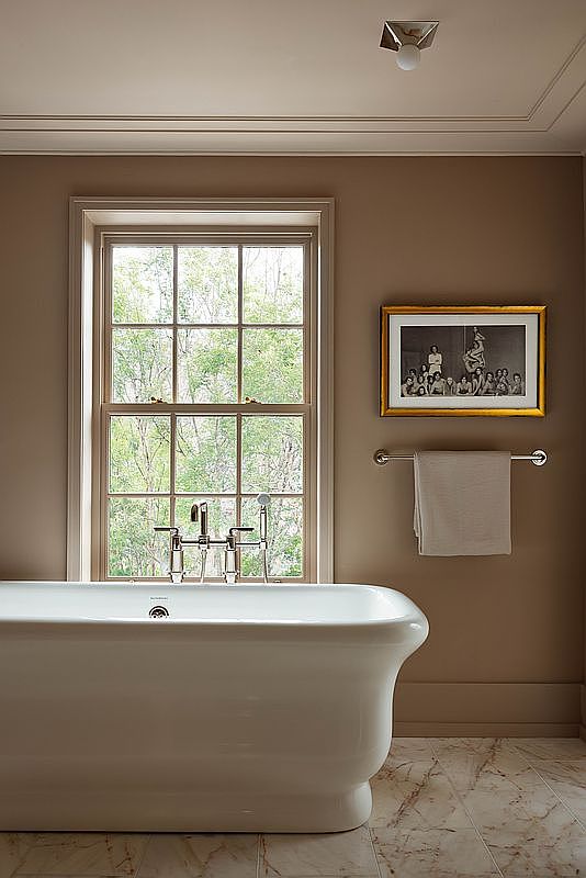 This elegant bathroom features a classic white freestanding soaking tub positioned beneath a large, multi-pane window that brings in natural light and views of the outdoors. The walls are painted in a sophisticated, warm taupe tone, complemented by a framed black-and-white photograph and a polished chrome towel rack. The space is finished with luxurious marble-patterned flooring, creating a serene and timeless atmosphere.
