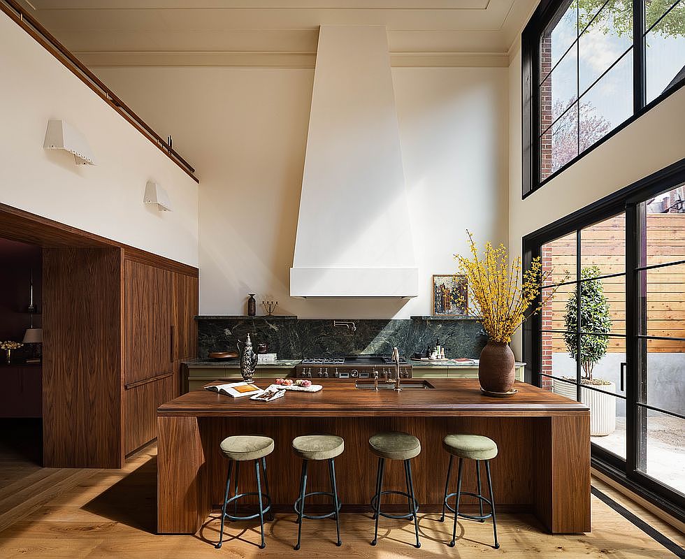 This sophisticated, high-ceilinged kitchen features a striking central island crafted from rich walnut wood, complemented by four velvet-upholstered stools. A dramatic, oversized white range hood serves as the focal point above a dark green marble backsplash and professional-grade cooktop. Large floor-to-ceiling windows flood the space with natural light, creating an airy, luxurious atmosphere that seamlessly blends modern design with warm, organic textures.