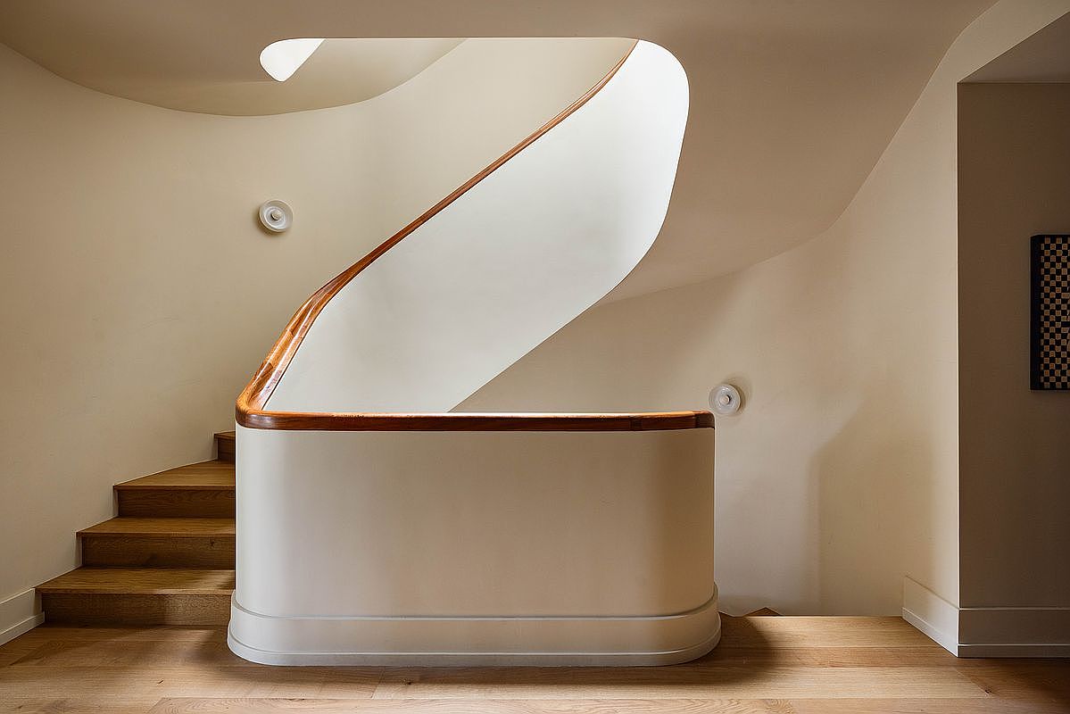 This elegant interior space features a striking, curved staircase with a warm wood handrail and matching wooden steps. The minimalist design is highlighted by smooth, off-white walls and a soft, diffused light that creates a serene and sophisticated atmosphere. The perspective captures the graceful flow of the architecture, emphasizing the clean lines and high-end craftsmanship of the home's transition area.