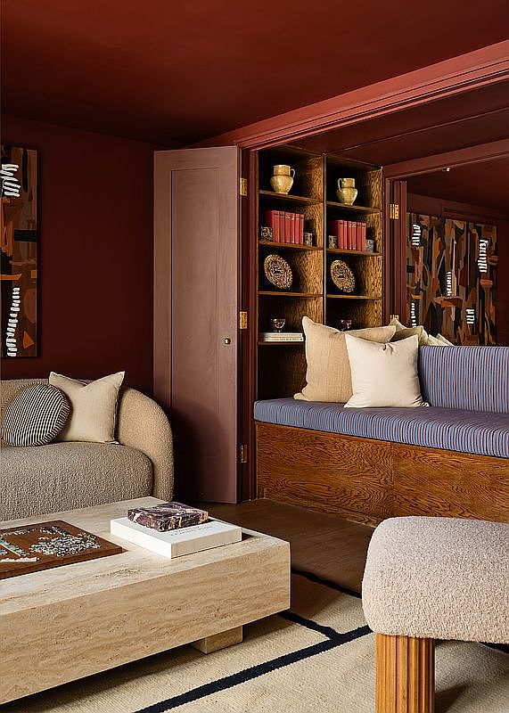 This sophisticated living room features a rich, monochromatic deep red color palette that creates a cozy and intimate atmosphere. The space includes a textured cream-colored sofa, a large travertine coffee table, and a built-in wooden bench seat with blue striped upholstery nestled against custom shelving. The room is accented by modern abstract art and a neutral rug with dark geometric lines, offering a blend of contemporary design and warm, inviting textures.