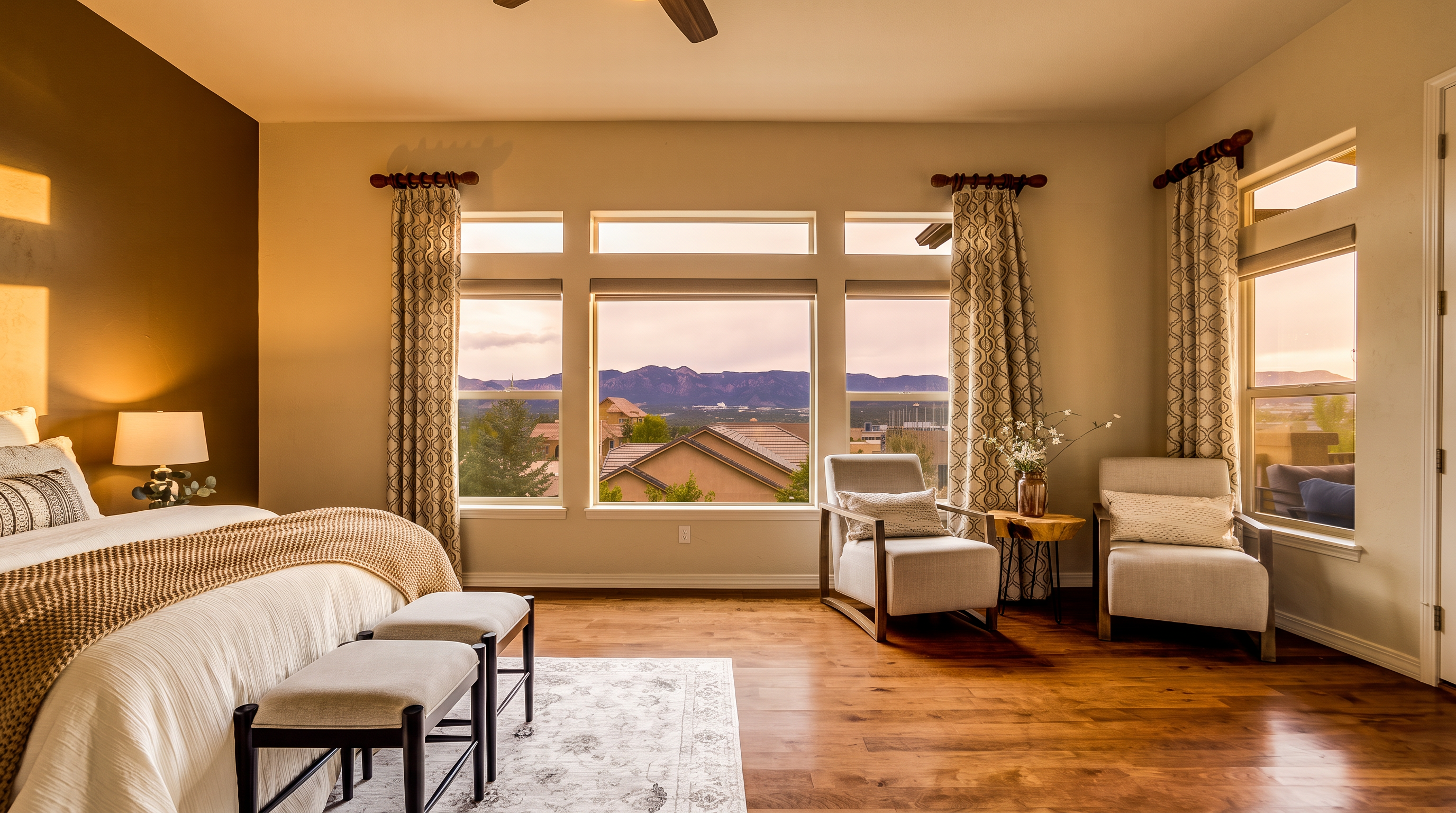 This spacious primary bedroom features warm wood flooring and a neutral color palette, centered around a large window arrangement that frames expansive mountain views. The room is furnished with a plush bed, two upholstered benches at the foot, and a cozy seating area with two armchairs and a small side table. The space is bright and airy, with patterned curtains adding a touch of texture to the serene, well-appointed interior.