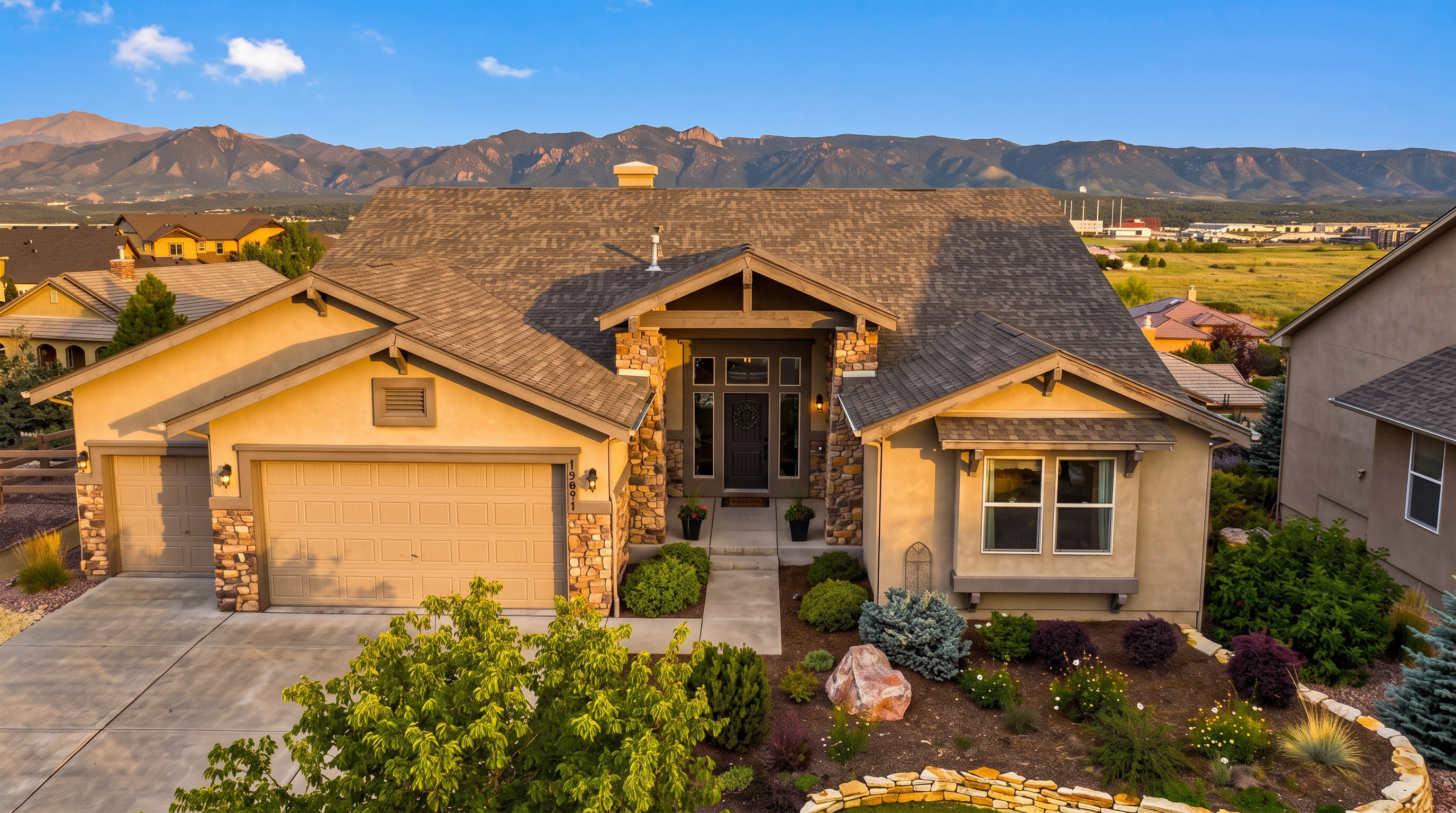 This elevated front-facing view captures a suburban home with a blend of stucco and stone facade, featuring a multi-gabled roofline and a prominent garage. The entryway is framed by stone pillars, leading to a dark-colored front door, while the front yard is landscaped with shrubs, a stone retaining wall, and a small tree. The backdrop showcases a scenic mountain range under a clear blue sky, emphasizing the property's desirable location.