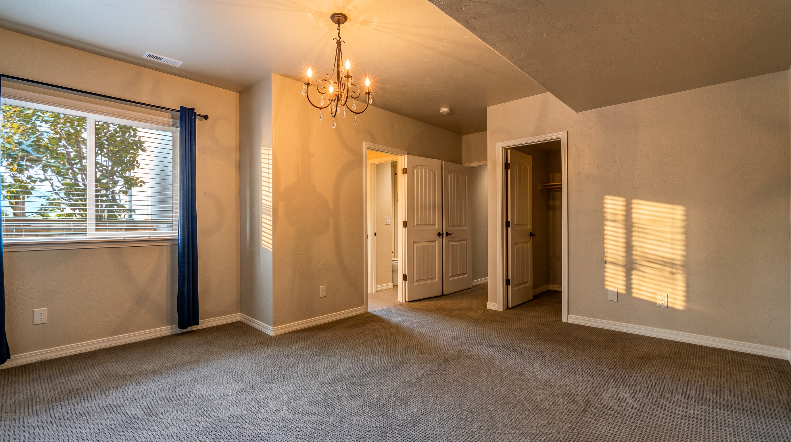 This bedroom features neutral-toned walls and wall-to-wall carpeting, creating a clean and versatile living space. A decorative chandelier hangs from the ceiling, while a window with dark curtains provides natural light. The room includes two doorways, one leading to an adjacent bathroom and the other to a walk-in closet, offering a functional layout.