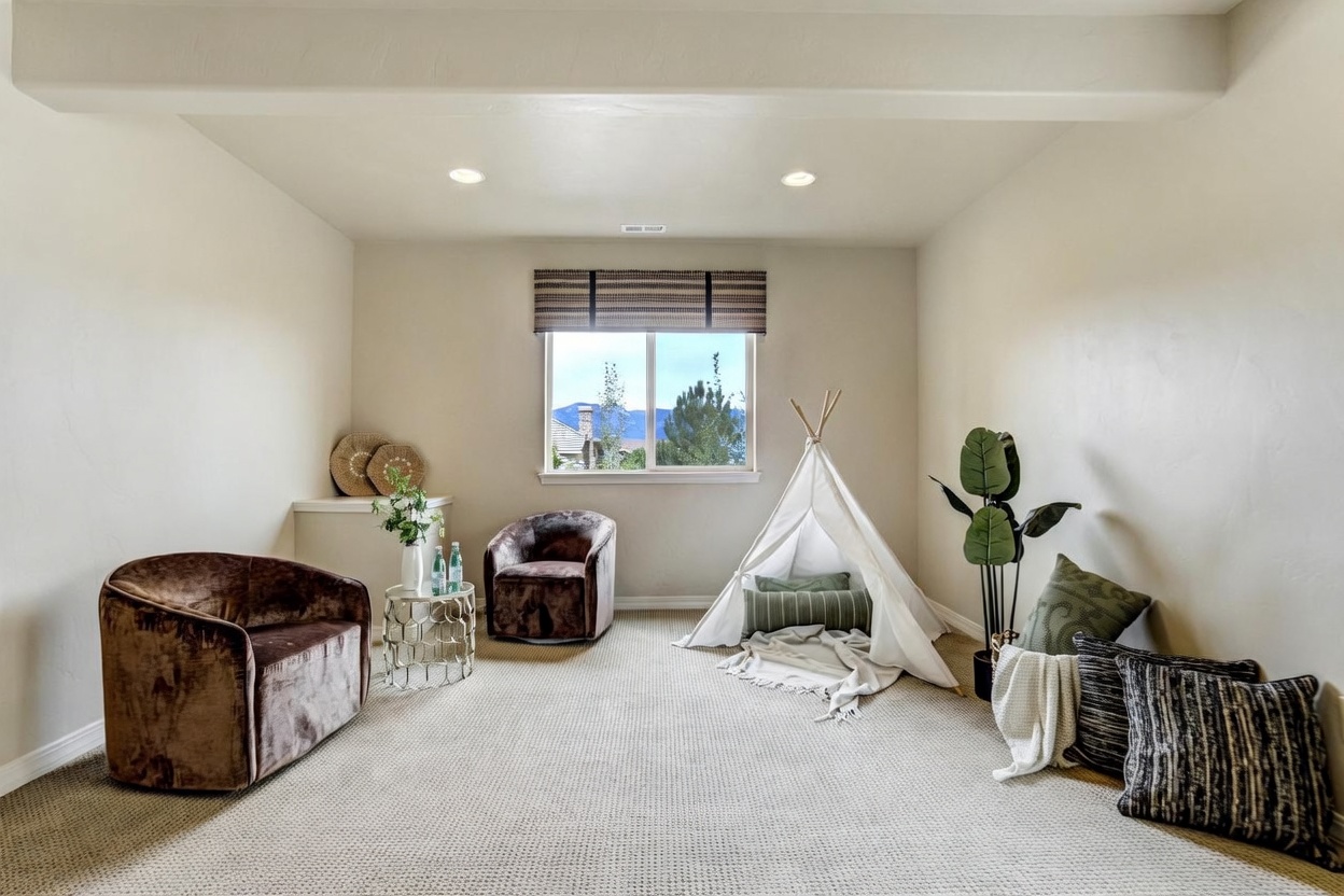 This cozy, carpeted room serves as a versatile play or lounge area, featuring two plush, dark brown velvet armchairs and a whimsical white fabric teepee. A small, round metallic side table with a decorative vase sits between the chairs, while a large potted plant and a collection of textured throw pillows add warmth to the corner. The space is illuminated by recessed lighting and a central window that provides a view of the outdoors, creating a bright and inviting atmosphere.