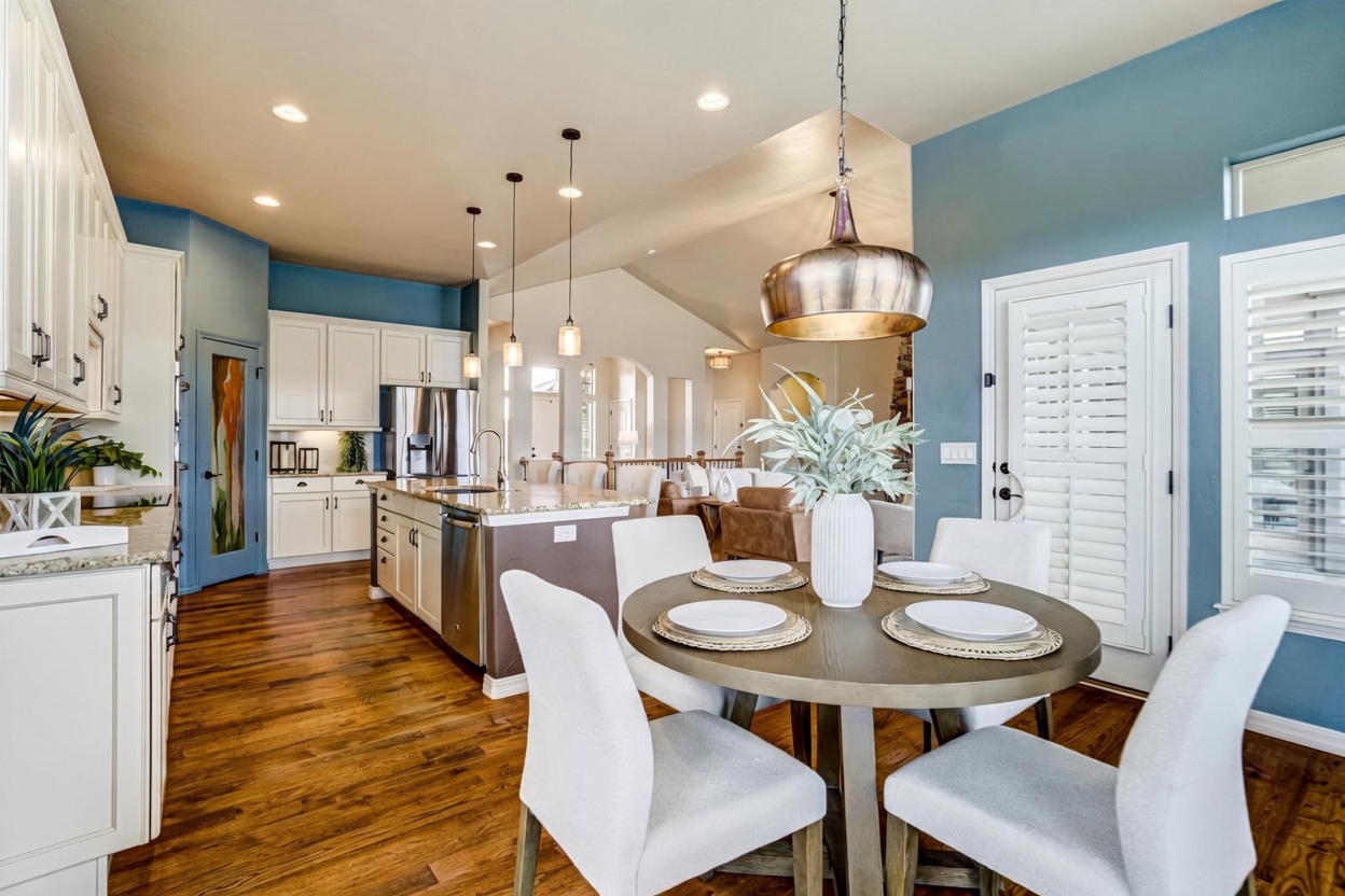 This open-concept kitchen and dining area features a central island with a stone countertop and bar seating, complemented by white cabinetry and stainless steel appliances. A round wooden dining table with four upholstered chairs sits in the foreground under a large metallic pendant light. The space is finished with warm wood flooring and blue accent walls, creating a bright and inviting atmosphere that flows into the adjacent living area.