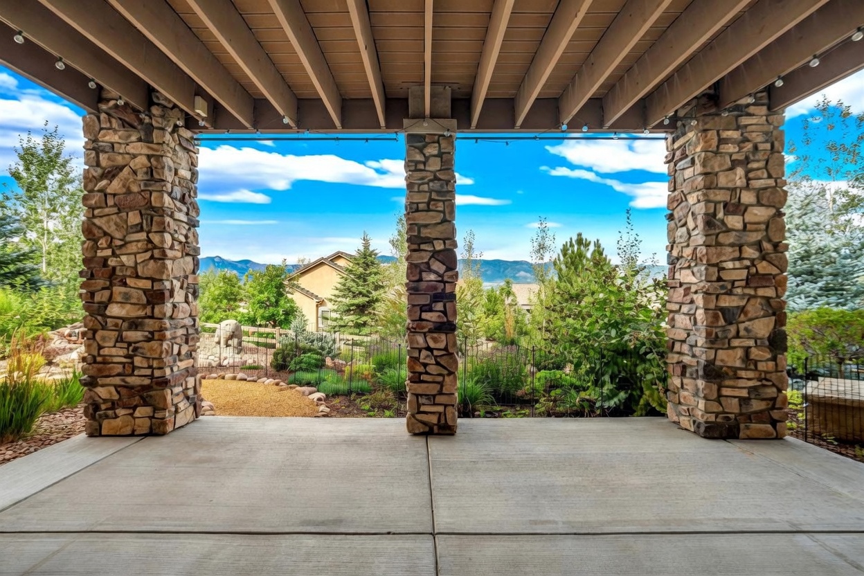 This covered patio features a concrete floor and three prominent stone-clad support columns that frame a scenic view of the surrounding landscape and distant mountains. The open-air design creates a seamless transition to the backyard, which includes a landscaped garden, a small gravel area, and mature trees. The perspective is from under the patio roof, looking outward toward the horizon, emphasizing the peaceful and expansive outdoor living space.