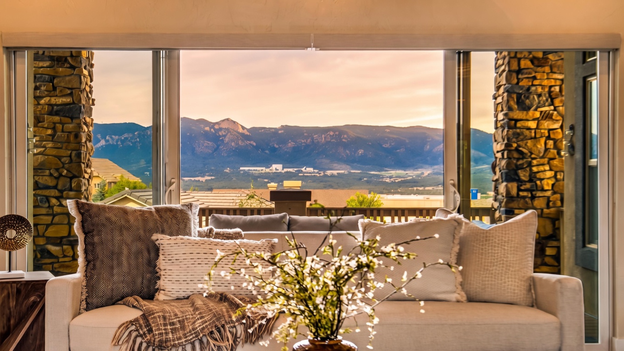 This interior living space features a comfortable sofa adorned with textured throw pillows and a draped blanket, positioned to face a expansive window wall. The view through the large glass panels showcases a scenic mountain range and a residential neighborhood below. Flanking the windows are rustic stone-clad pillars, adding a natural, textured element to the room's design.