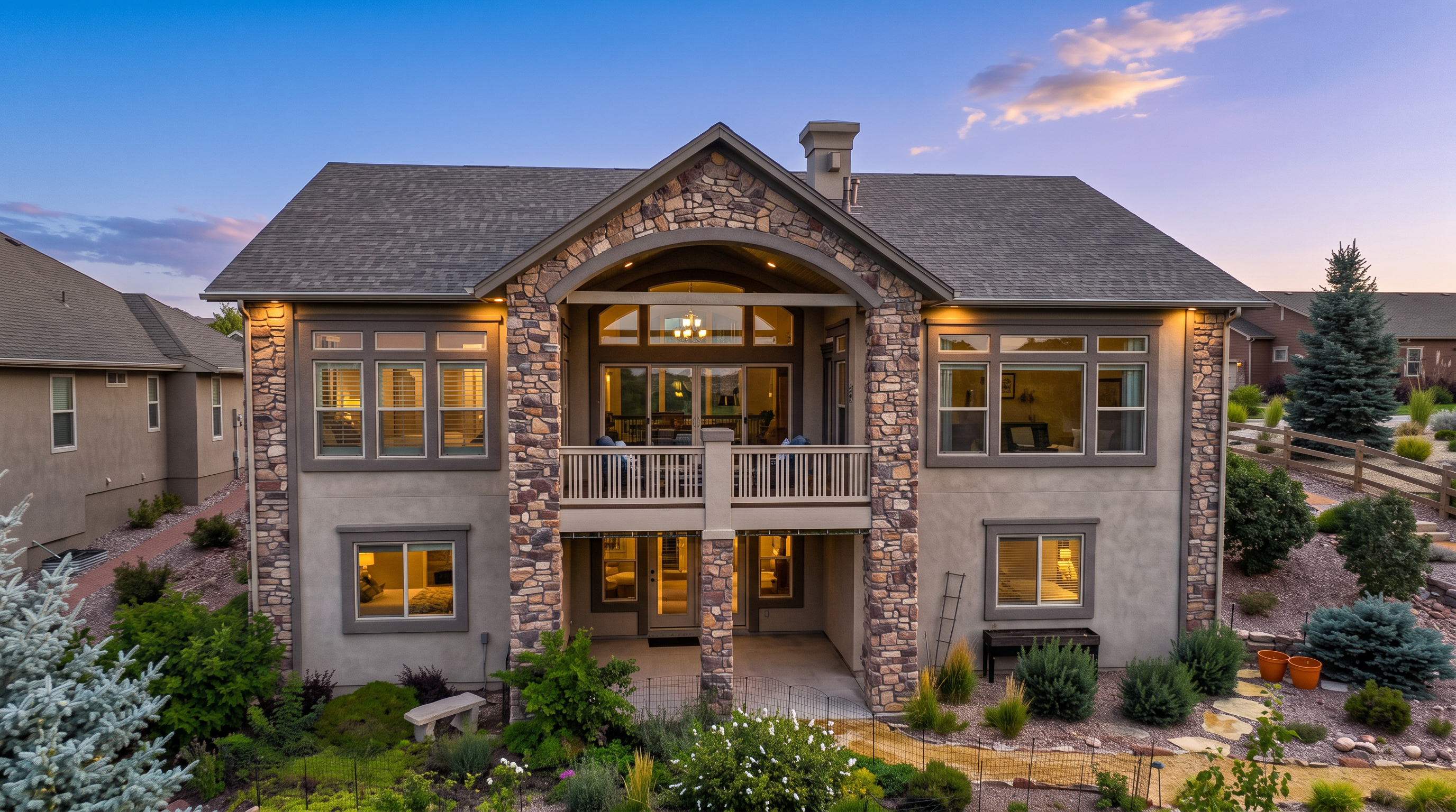 This rear view of a multi-story suburban home showcases a prominent stone-clad central section featuring a covered upper-level balcony and a ground-level patio. The exterior is finished in a neutral stucco with stone accents, complemented by a multi-gabled roofline and large windows that provide ample natural light. The property includes a landscaped backyard with a stone retaining wall, a winding gravel path, and mature evergreen trees, creating a serene and well-maintained outdoor living space.