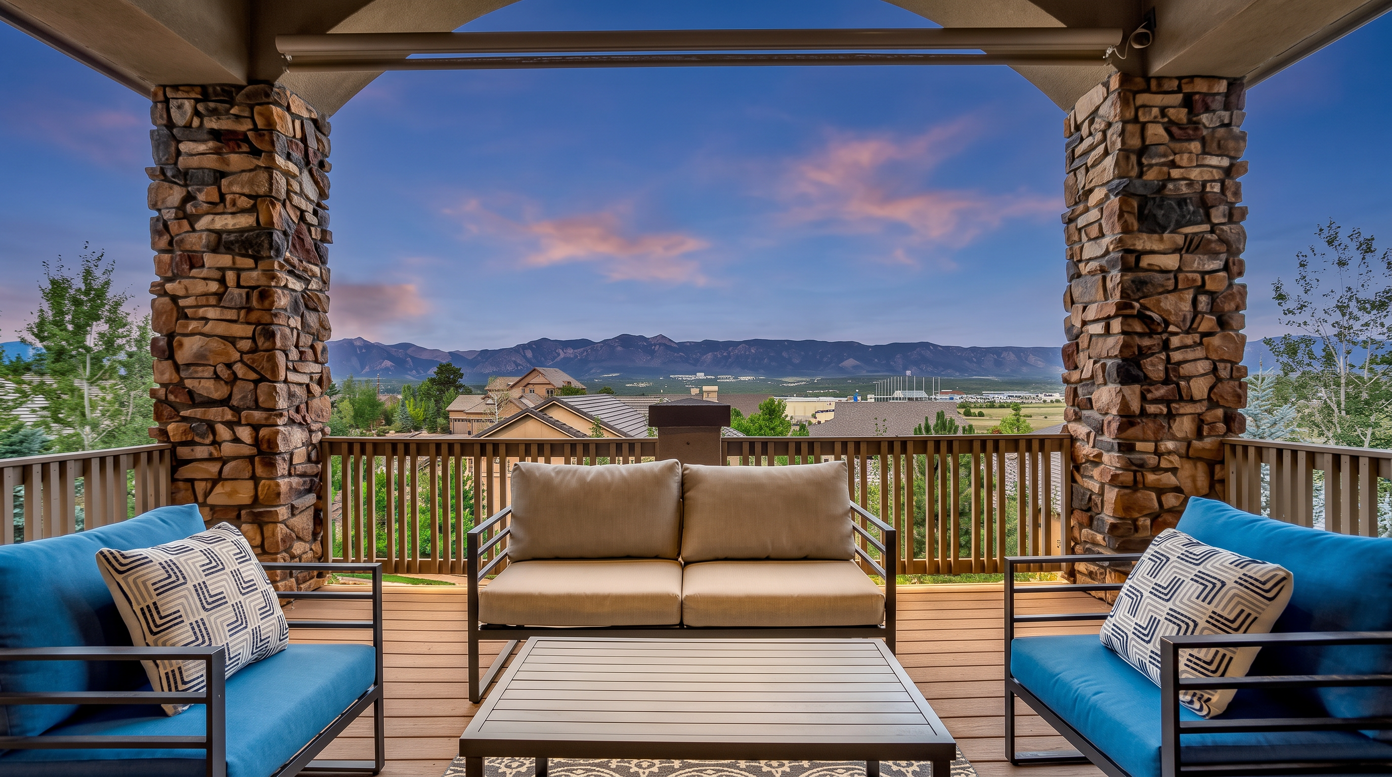 This elevated balcony offers a stunning, unobstructed view of distant mountain ranges under a bright blue sky. The space is furnished with a comfortable outdoor seating arrangement, including a central sofa and two matching armchairs with blue cushions, all set upon a patterned area rug. The balcony is framed by sturdy stone-clad pillars and a wooden railing, creating a rustic yet refined outdoor living area perfect for relaxation.
