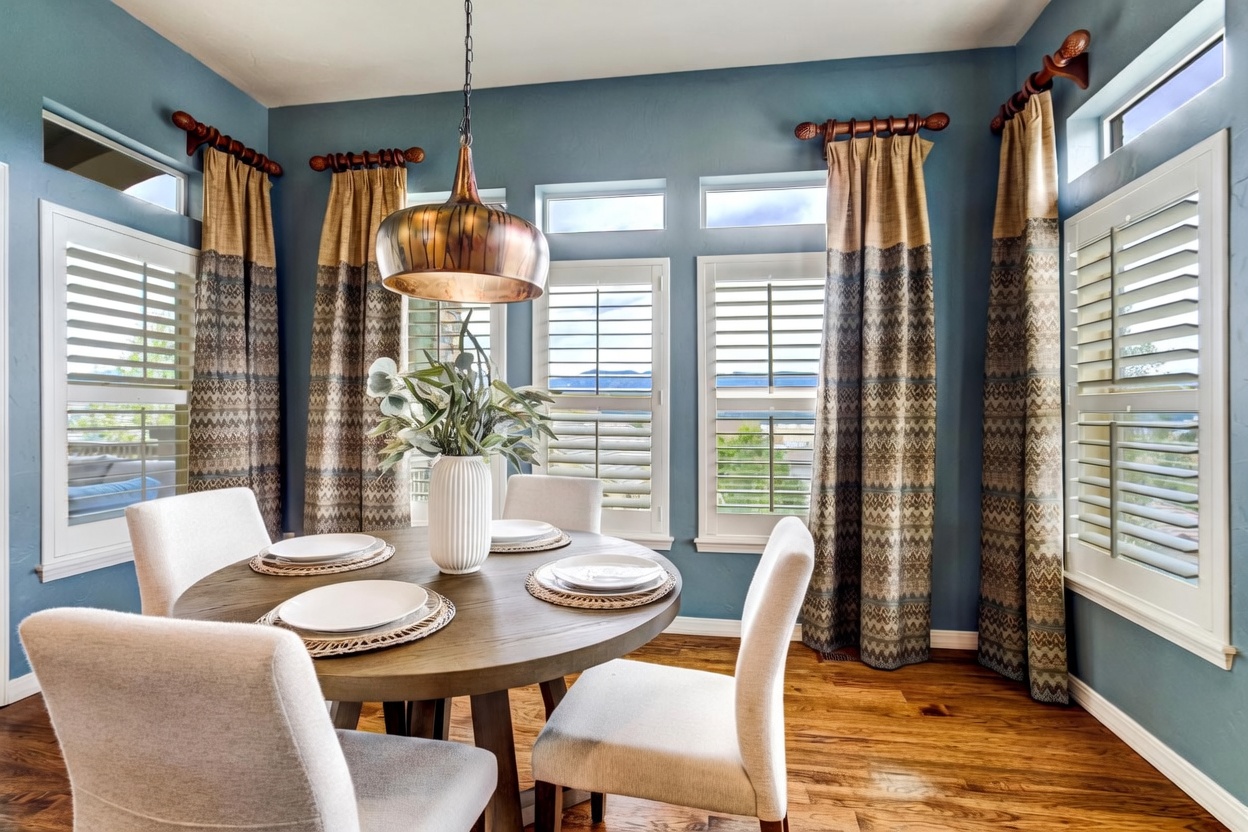 This bright dining area features a round wooden table set for four, surrounded by light-colored upholstered chairs. The room is defined by deep blue walls, tall windows with white shutters, and patterned floor-length curtains that add texture to the space. A large, metallic pendant light hangs above the table, which is centered on a warm-toned wood floor, creating an inviting and sophisticated atmosphere.
