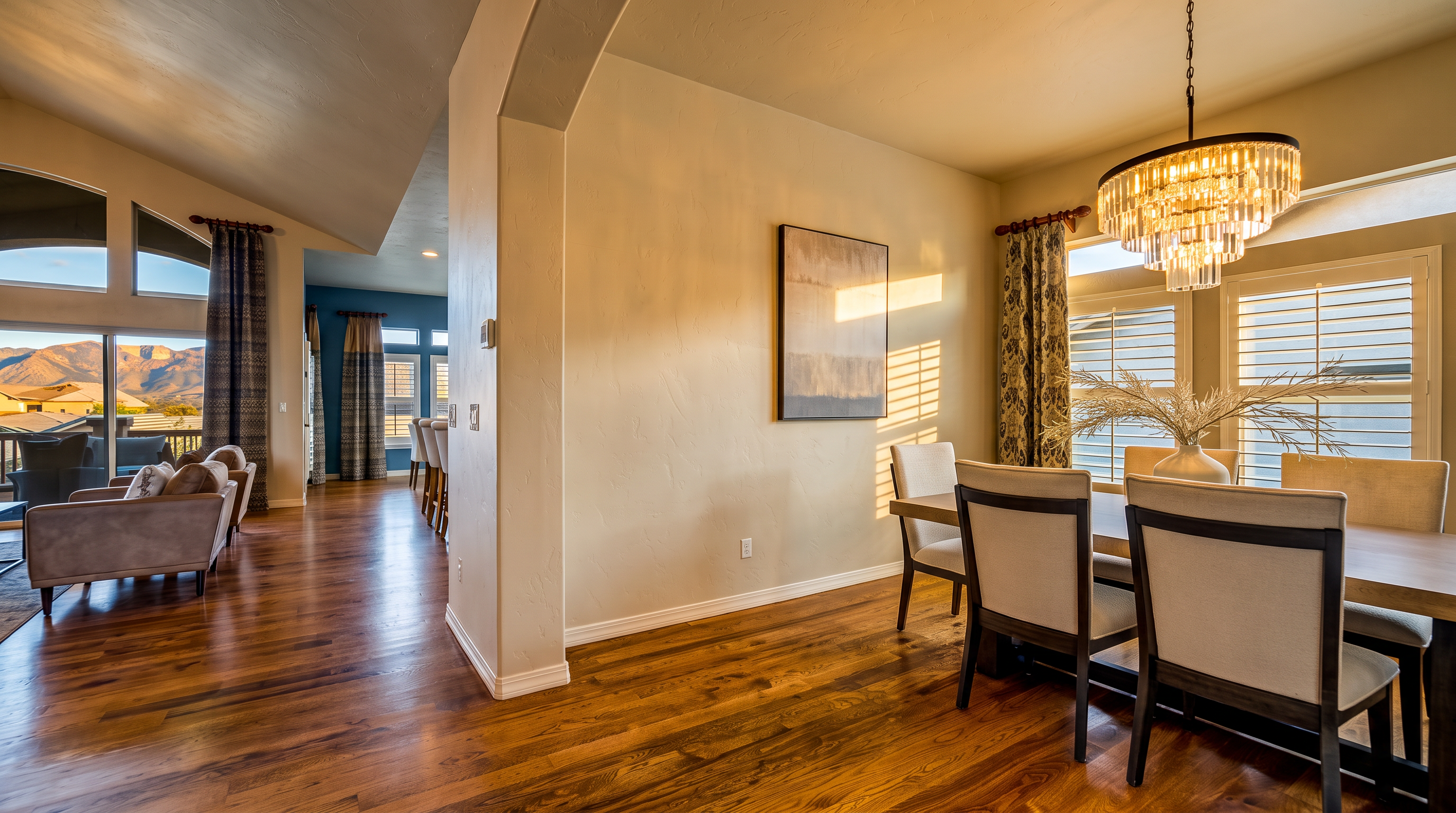 This dining area features a sophisticated, open-concept design with rich hardwood flooring and a prominent crystal chandelier as a focal point. The space includes a dining table with upholstered chairs and is framed by tall windows dressed in patterned curtains, offering a view of the distant mountains. A neutral-toned abstract piece of art hangs on the wall, bridging the transition between the dining area and the adjacent living space.