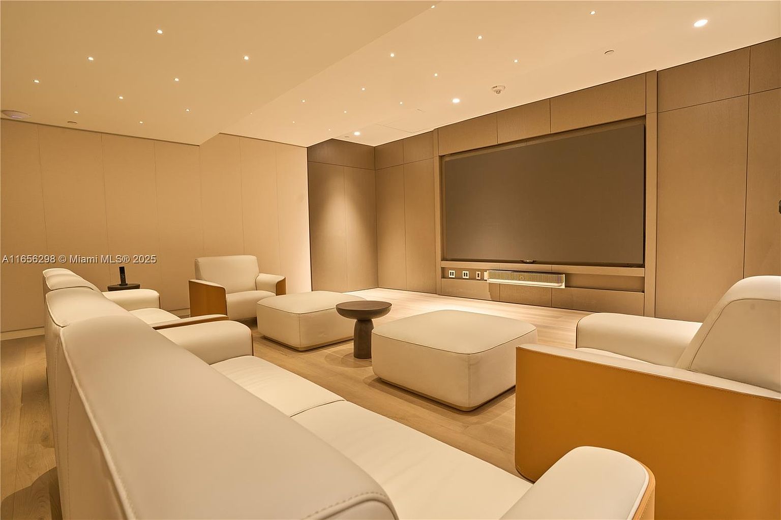 This is a modern living room featuring a large sectional sofa, two armchairs, and two ottomans, all in a light neutral color. A large flat-screen television is mounted on a paneled wall, and a small round table sits between the ottomans. The room has a minimalist aesthetic with recessed lighting and light wood flooring, creating a clean and inviting space.