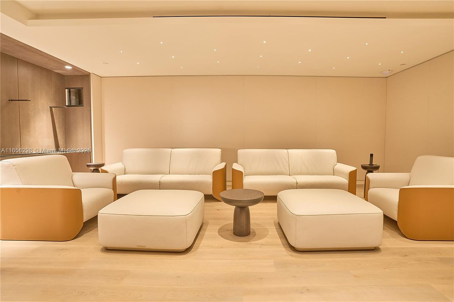 This interior shot showcases a modern living room with a minimalist design. The room features two cream-colored sofas, two matching armchairs with tan accents, and two square ottomans, arranged around a small, round coffee table. The light wood flooring and recessed lighting contribute to a bright and inviting atmosphere, perfect for a sophisticated and comfortable living space.