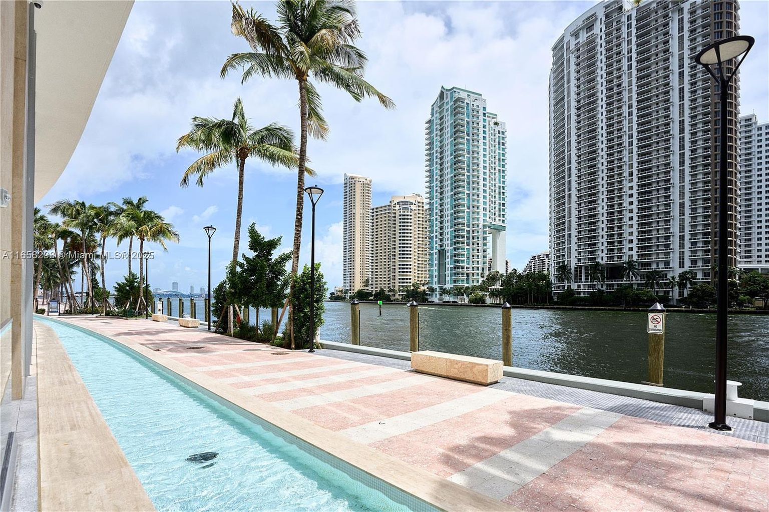 This image showcases a luxurious outdoor pool area alongside a waterfront promenade. The scene features modern high-rise buildings in the background, palm trees lining the walkway, and a narrow, elegant pool running parallel to the pedestrian path. The overall impression is one of upscale urban living with a focus on leisure and scenic views.