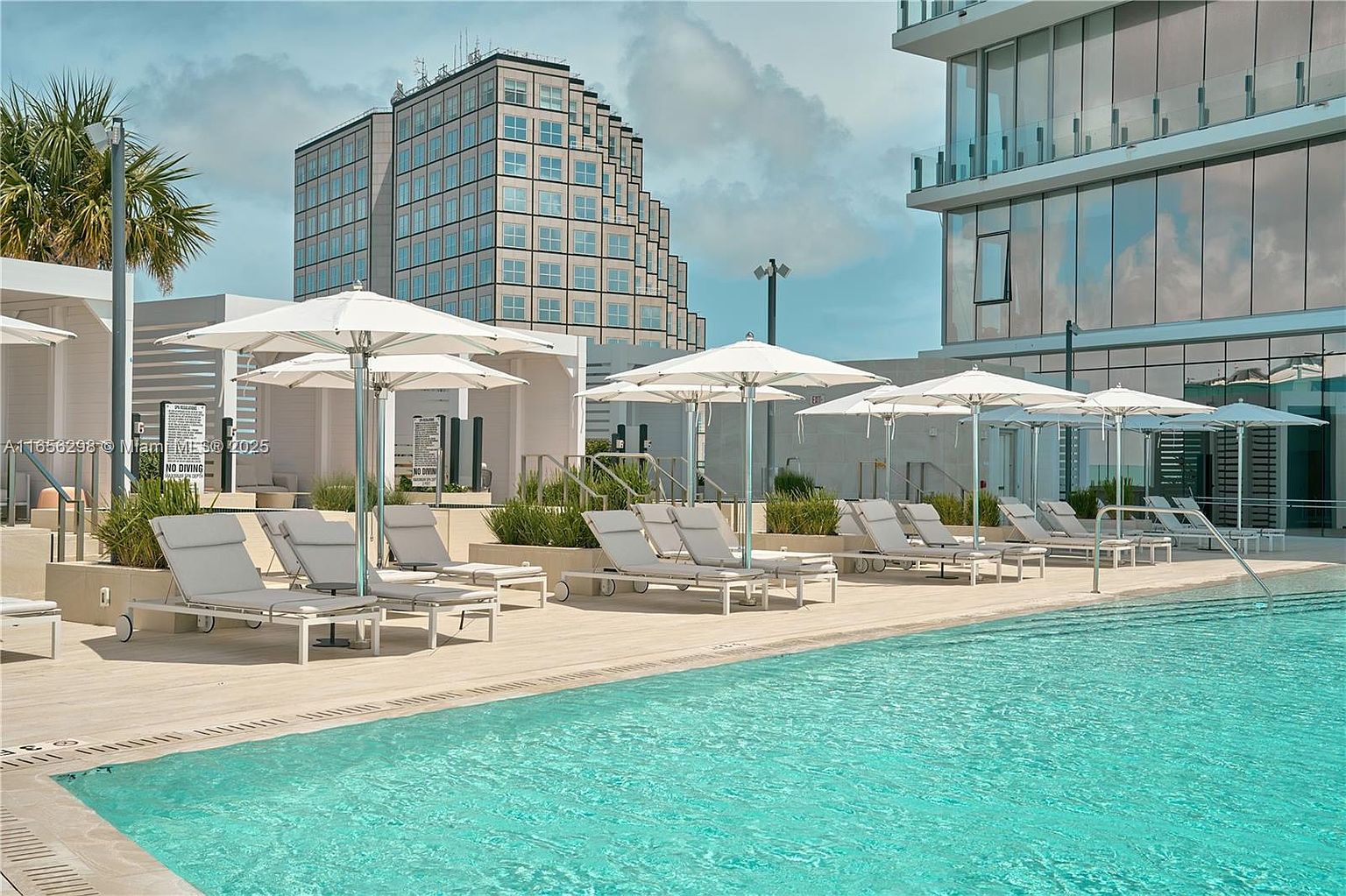 This image showcases a luxurious outdoor pool area, complete with modern lounge chairs and large white umbrellas for shade. The pool's turquoise water contrasts beautifully with the light-colored decking, while contemporary buildings rise in the background, suggesting an upscale residential complex. The scene evokes a sense of relaxation and high-end living.