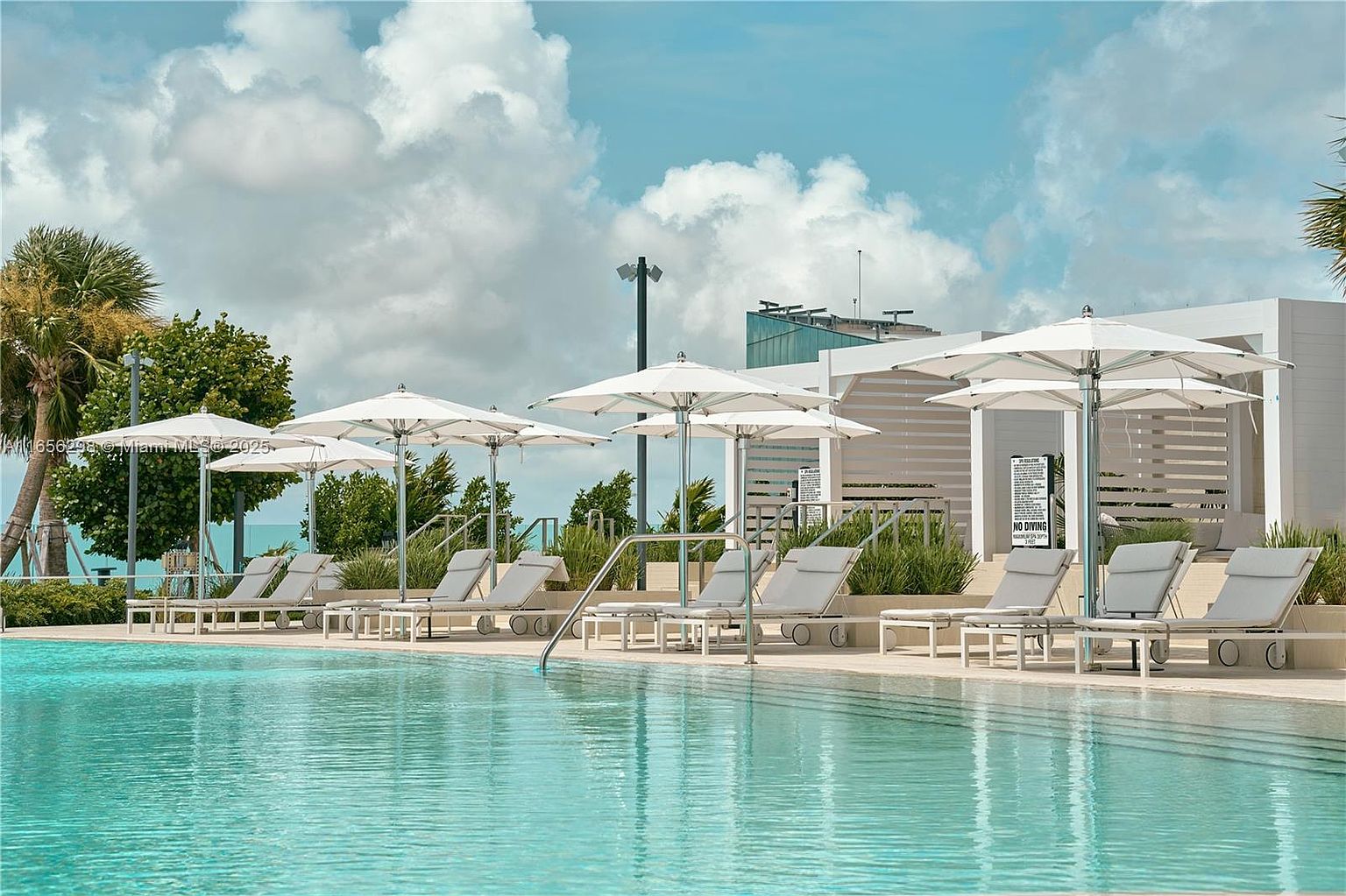 The image showcases a luxurious pool area with several lounge chairs and white umbrellas arranged neatly along the pool's edge. The turquoise water of the pool reflects the sky, creating a serene and inviting atmosphere. The scene is framed by lush greenery and modern architectural elements, suggesting a high-end resort or residential complex.