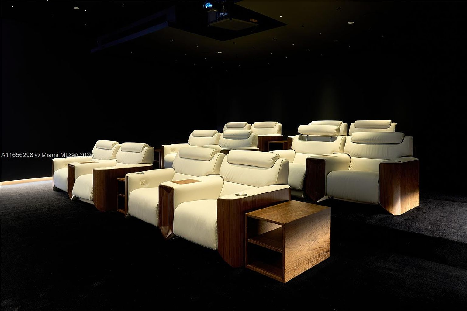 This is an interior shot of a home theater. The room features multiple rows of plush, white leather reclining seats with wooden side tables and cup holders. The ceiling is dark with small, scattered lights, creating a starry effect, and a projector is mounted on the ceiling.
