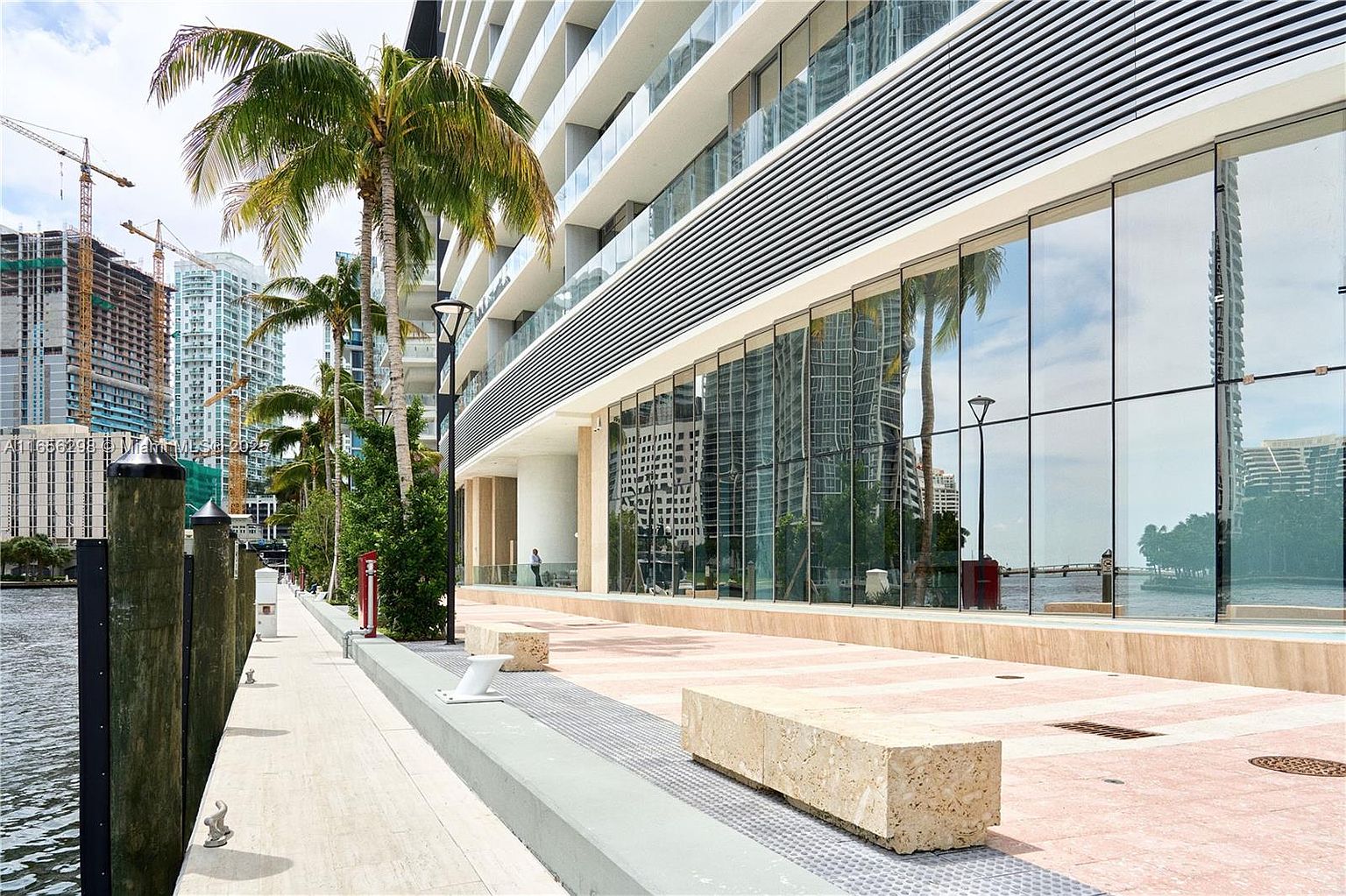 This image showcases the side exterior of a modern building, likely a condominium or apartment complex, situated along a waterfront. The building features a sleek design with large glass windows reflecting the surrounding cityscape and water. A pedestrian walkway with stone benches runs alongside the building, enhanced by palm trees and modern lighting, creating an inviting and upscale atmosphere.