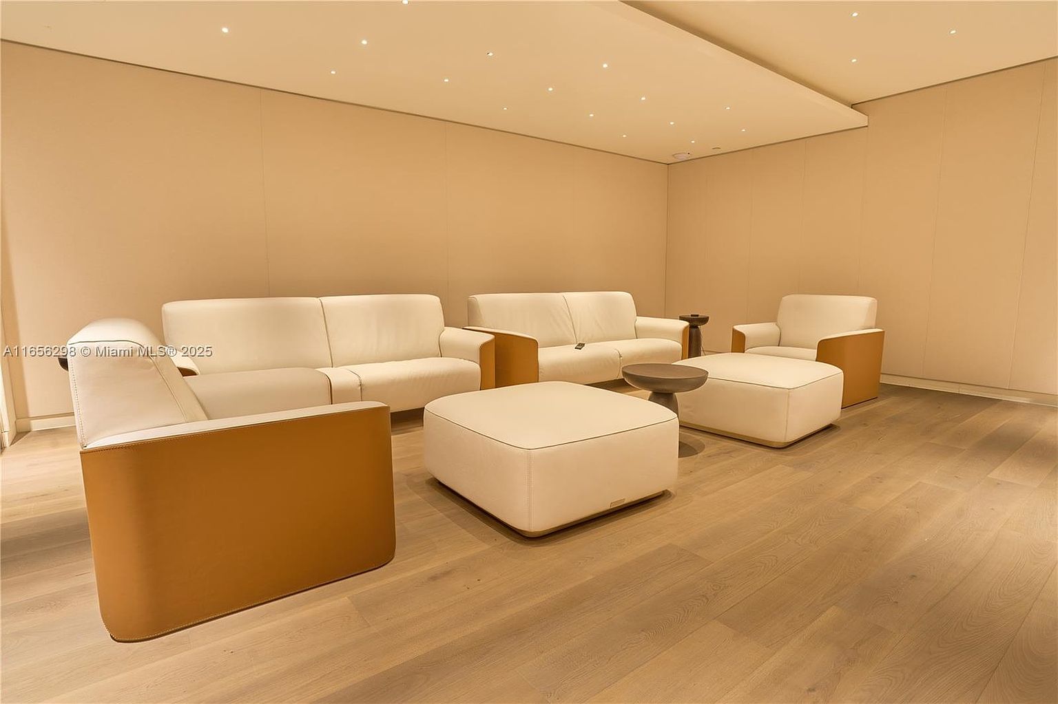 This interior shot showcases a modern living room with neutral tones. The room features two white sofas, an armchair, and ottomans, all arranged on a light wood floor. The walls are a soft beige, and recessed lighting provides a clean, minimalist aesthetic, creating a serene and inviting atmosphere.