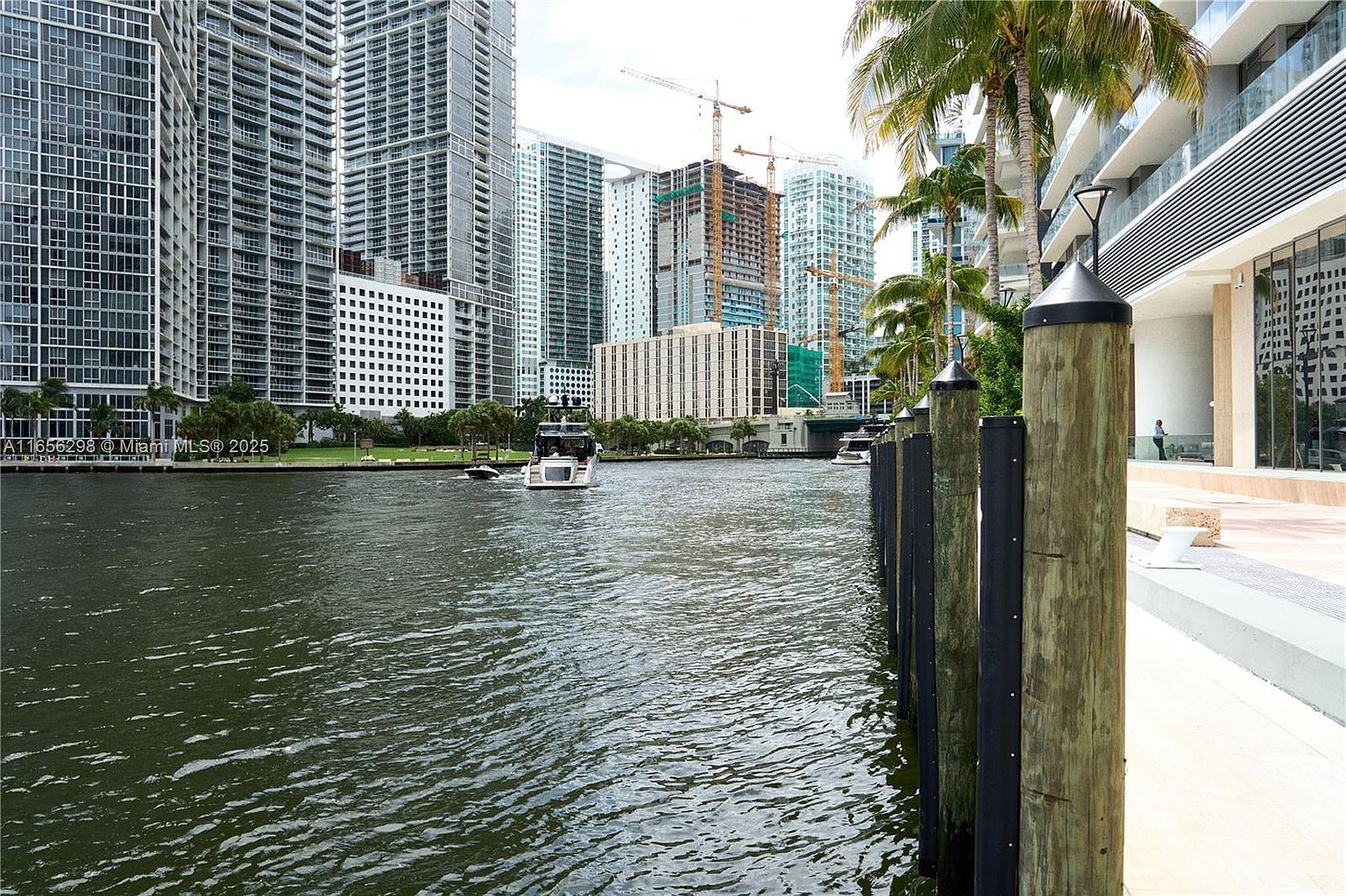 This exterior shot showcases a waterfront view with modern high-rise buildings in the background. The water is calm, reflecting the cityscape, and several boats are visible. Wooden pylons line the edge of a walkway, adding a nautical touch to the scene.