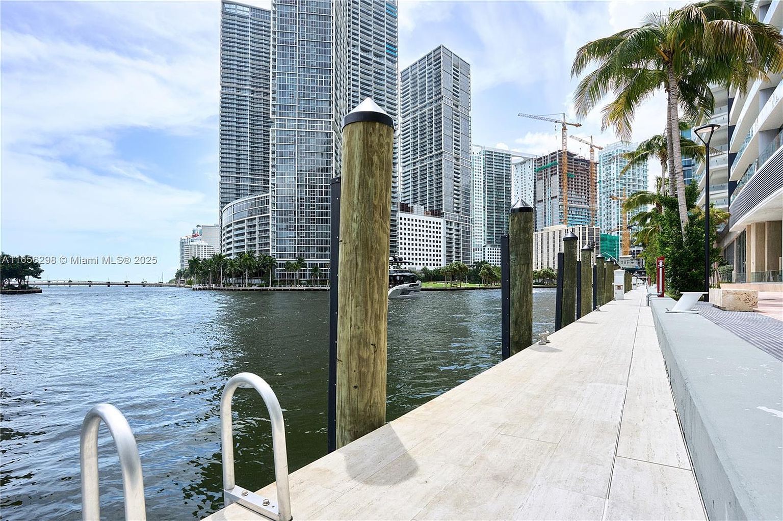 This image showcases a modern waterfront patio or deck area, featuring sleek, light-colored paving stones and wooden pilings along the water's edge. In the background, towering high-rise buildings create a dramatic urban backdrop, while palm trees add a touch of tropical elegance. The scene suggests a luxurious and desirable waterfront property.