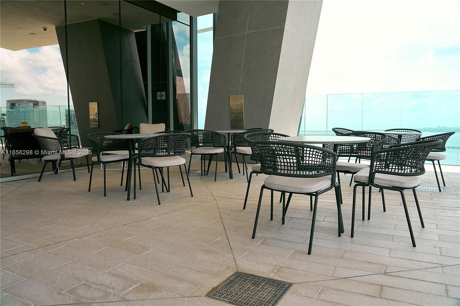 This image showcases a stylish outdoor patio or balcony area, furnished with modern black wicker chairs and round tables. The space features a sleek, contemporary design with a neutral color palette, complemented by the surrounding architecture and a glimpse of the water in the background. The area appears to be part of a high-end residential building, offering a sophisticated outdoor living experience.