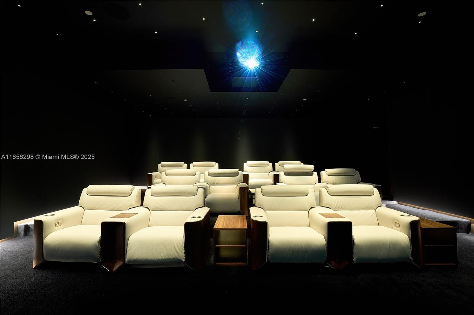 This is a luxurious home theater featuring multiple rows of plush, cream-colored reclining seats with dark wood accents and integrated cup holders. A bright projector beam illuminates the room from the ceiling, creating a cinematic atmosphere. The walls and ceiling are dark, enhancing the viewing experience and creating a sense of immersion.