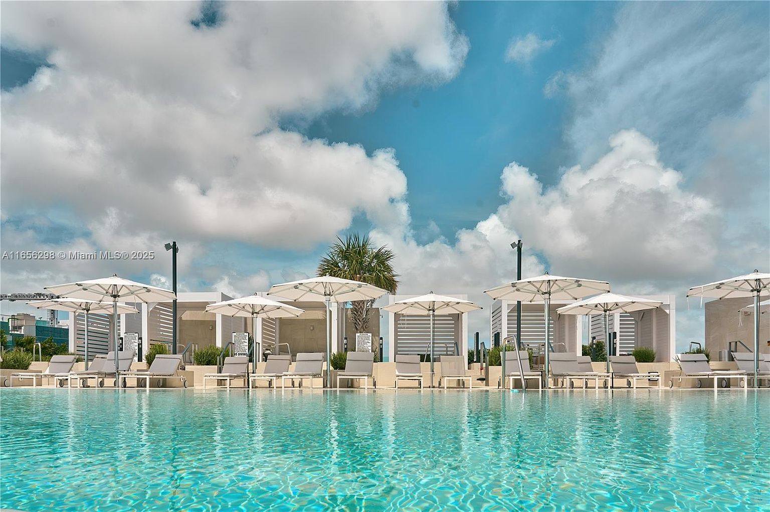 This image showcases a luxurious pool area with clear turquoise water, surrounded by elegant white cabanas and lounge chairs. The scene is set against a backdrop of a partly cloudy blue sky, creating a serene and inviting atmosphere. The composition emphasizes the upscale amenities and the potential for relaxation and leisure.
