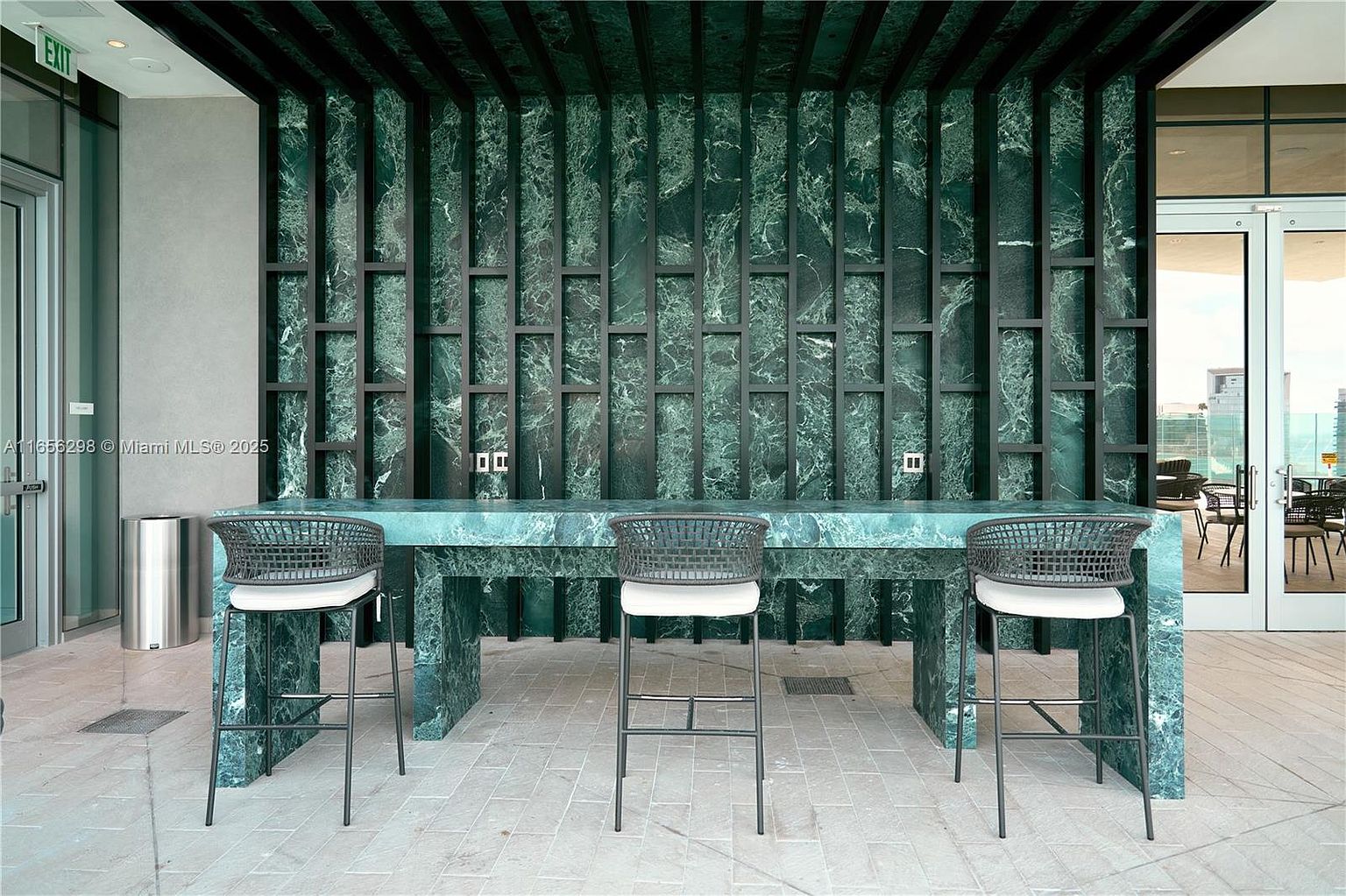 This image showcases a stylish outdoor patio or balcony area, featuring a unique green marble bar with integrated electrical outlets and three modern bar stools. The backdrop is a decorative green marble wall with a geometric black frame, adding a touch of sophistication. The space appears to be part of a luxury building, offering a chic and functional outdoor amenity.