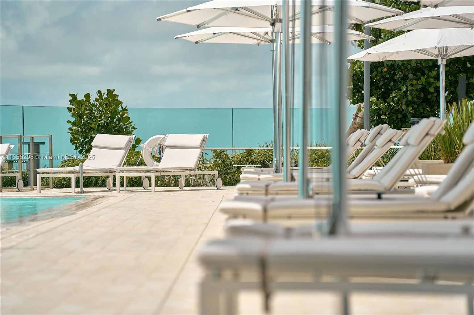 This image showcases a luxurious pool area with multiple white lounge chairs and matching umbrellas, creating a serene and upscale atmosphere. The pool is partially visible on the left, and a glass barrier provides a clear view of the sky and distant greenery. The scene evokes a sense of relaxation and high-end living, perfect for attracting potential buyers seeking a resort-style amenity.