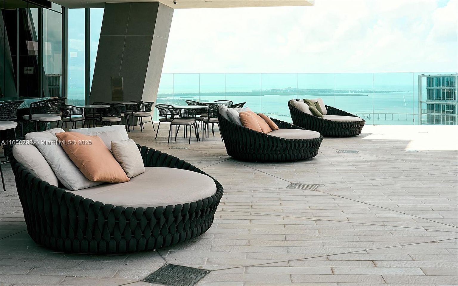 This image showcases a luxurious outdoor patio or balcony area, featuring stylish round lounge chairs with plush cushions, offering a comfortable seating arrangement. The space is designed with modern aesthetics, complemented by the sleek outdoor furniture and the stunning view of the ocean or bay in the background. The setting suggests a high-end residential property with an emphasis on relaxation and outdoor living.