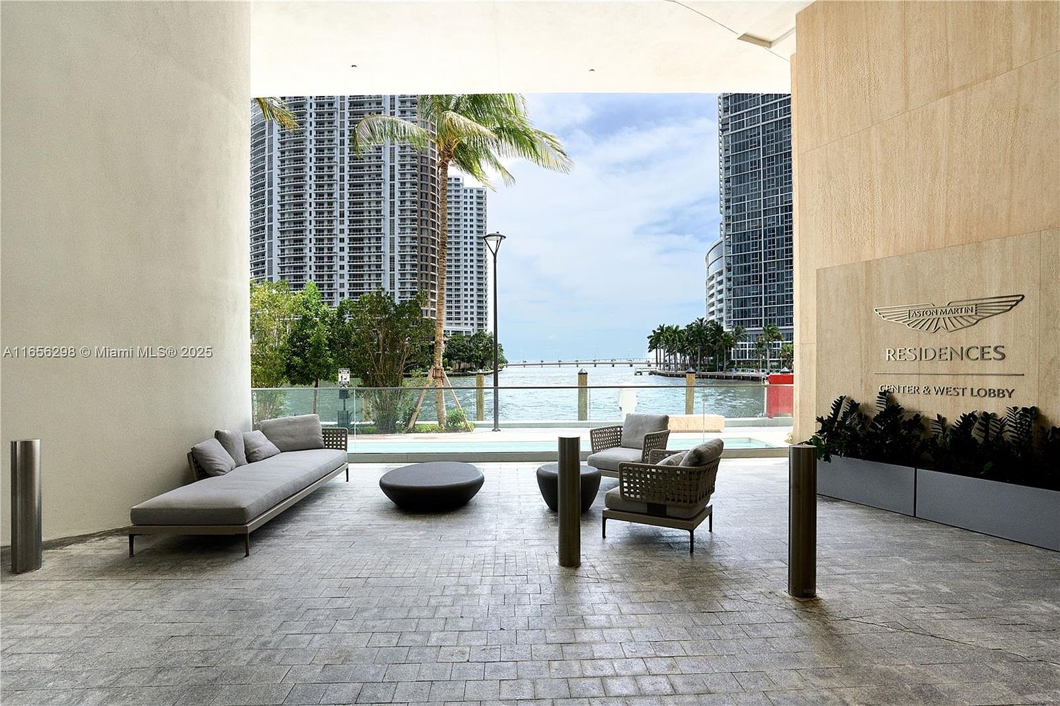 This image showcases a luxurious patio or balcony area with modern outdoor furniture, including a chaise lounge and two armchairs arranged around a low coffee table. The space offers stunning waterfront views framed by high-rise buildings, creating an upscale urban oasis. The Aston Martin Residences logo is visible, indicating the building's prestigious branding.