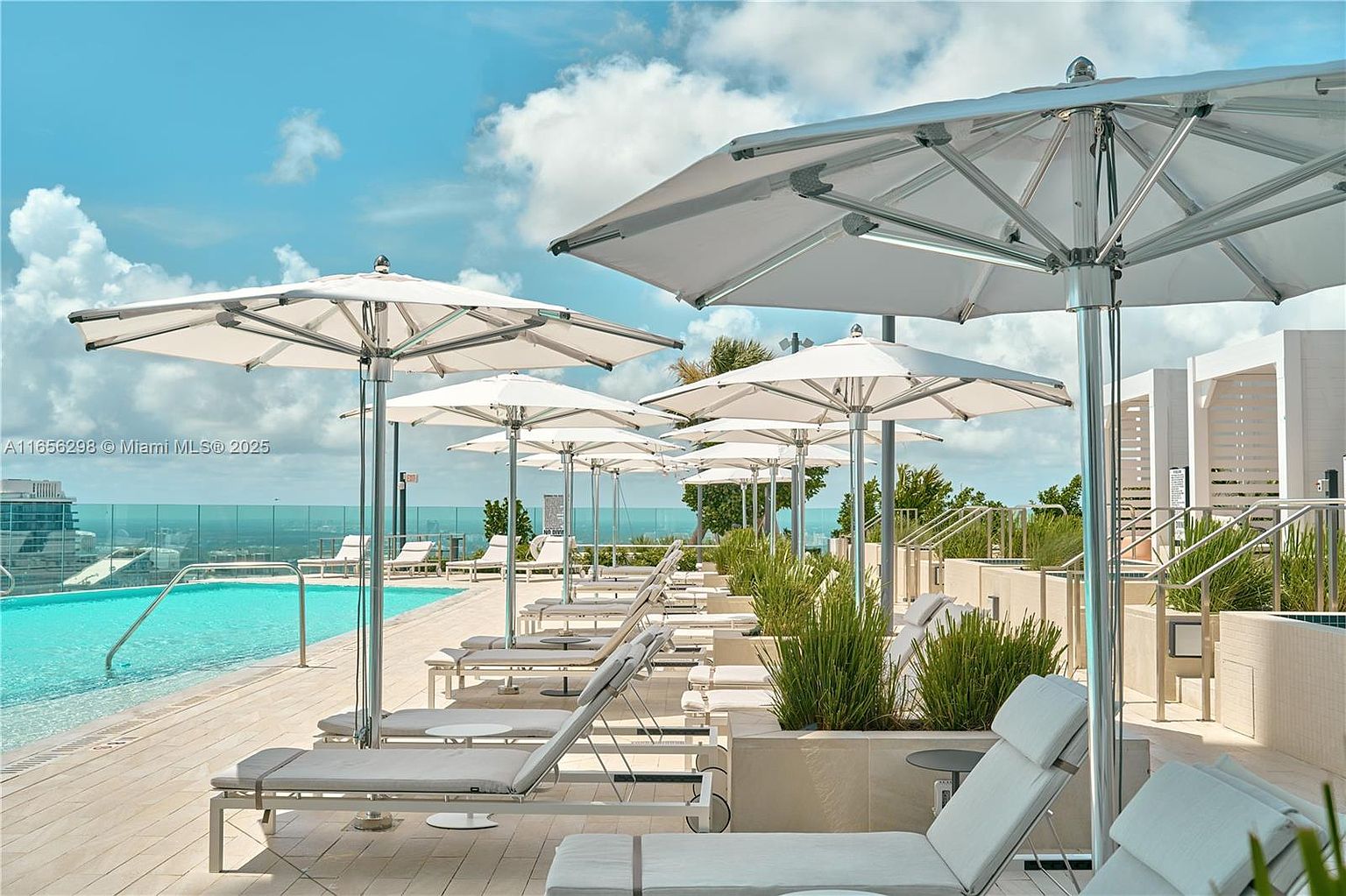 This image showcases a luxurious pool area with multiple lounge chairs and large white umbrellas providing shade. The turquoise pool water contrasts beautifully with the light-colored decking and the blue sky dotted with clouds. The scene evokes a sense of relaxation and upscale resort living, perfect for attracting potential buyers seeking a high-end lifestyle.