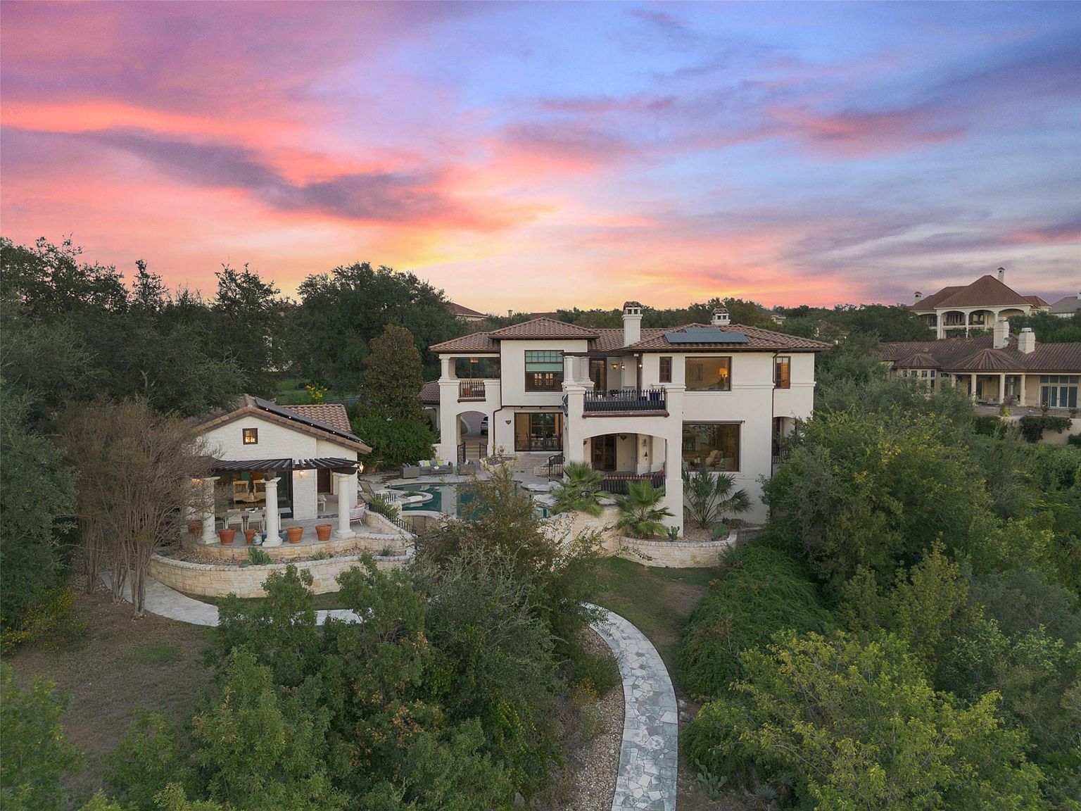 This aerial view showcases a luxurious estate at sunset, featuring a grand two-story Mediterranean-style home with a tile roof, balconies, and a swimming pool. A separate pool house with a pergola adds to the property's appeal, while a stone pathway winds through lush landscaping, creating a serene and private setting. The overall impression is one of elegance and exclusivity.