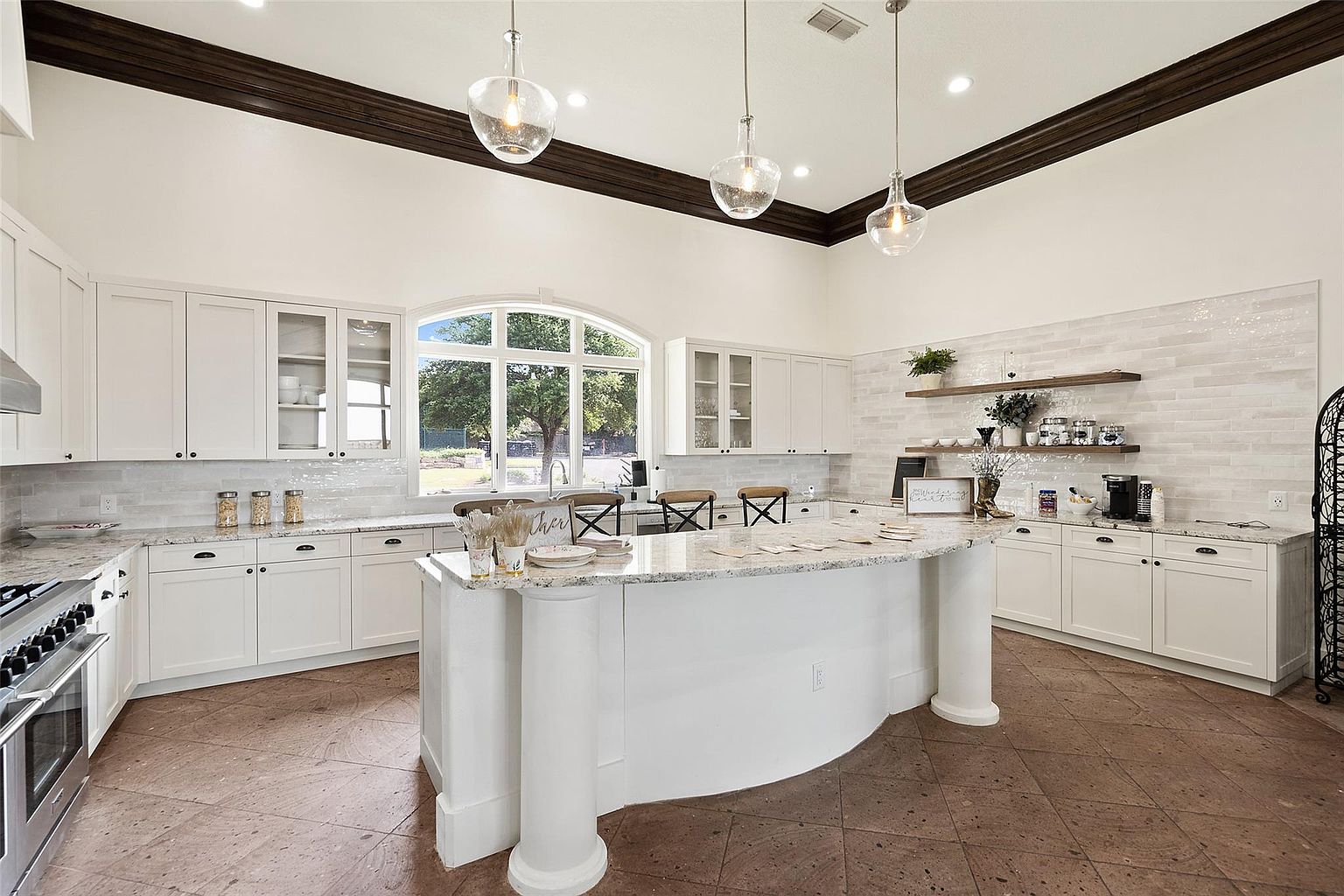 This is a bright and spacious kitchen featuring white cabinetry, granite countertops, and stainless steel appliances. A large island with decorative columns serves as a focal point, complemented by pendant lighting and a large arched window that provides natural light. The kitchen also includes open shelving and a decorative backsplash, creating a blend of functionality and style.