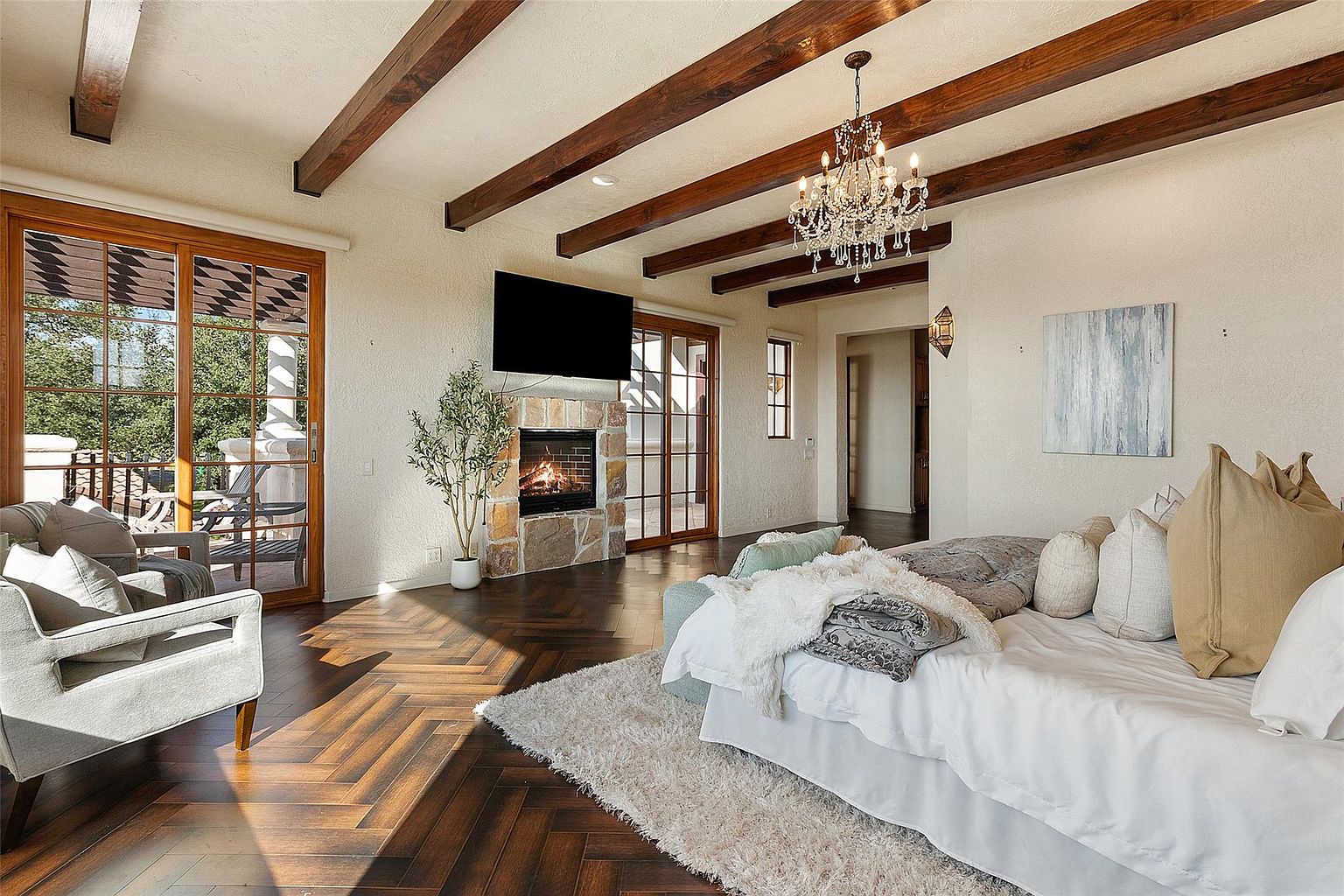 This is a primary bedroom featuring a stone fireplace with a mounted television above it, wooden ceiling beams, and a crystal chandelier. The room has a luxurious feel with a large bed covered in plush blankets and pillows, and a cozy armchair near the sliding glass doors that lead to a balcony. The dark wood flooring adds warmth and elegance to the space.