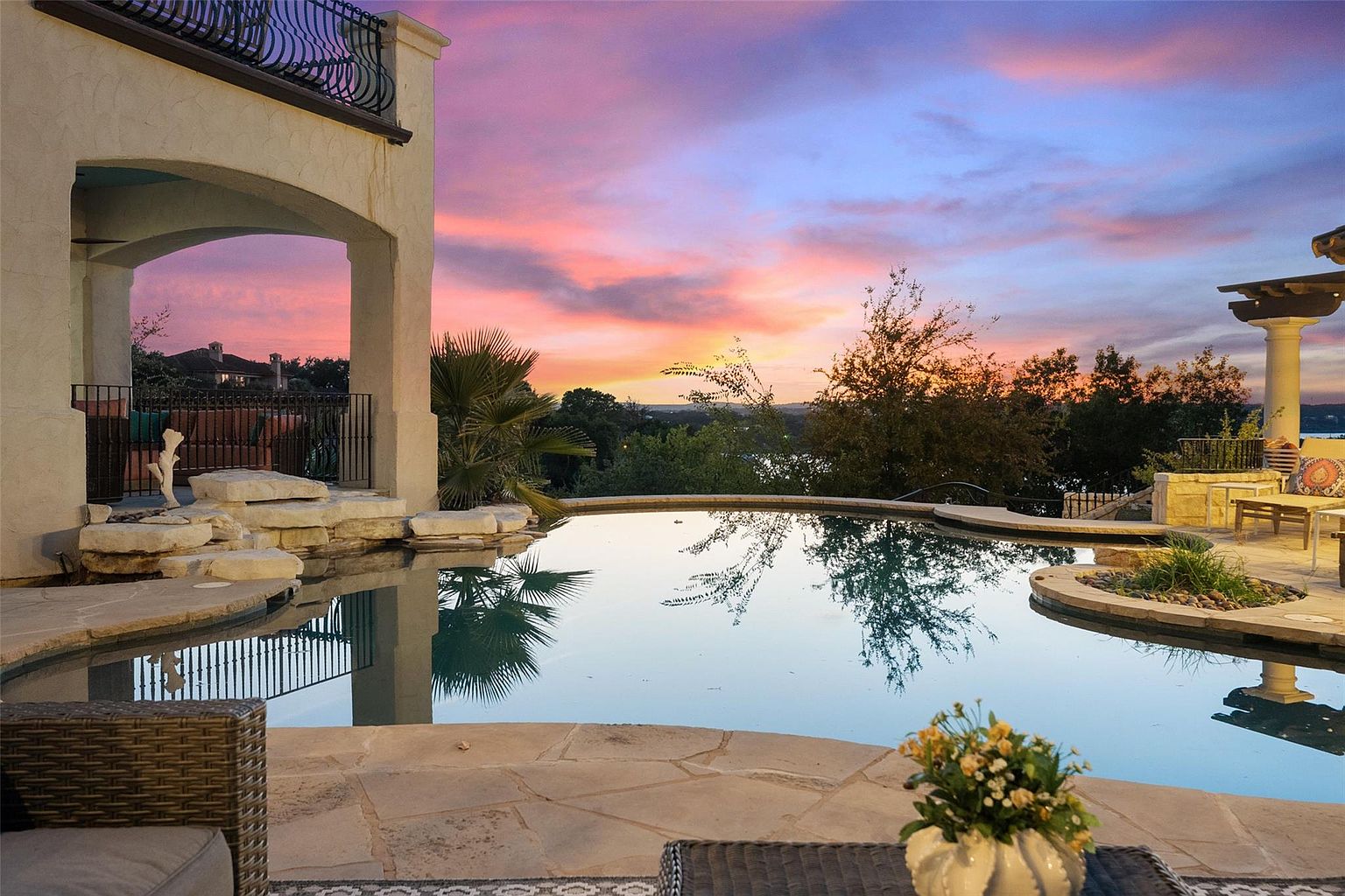 This image showcases a luxurious outdoor pool area at sunset, featuring a freeform pool with integrated spa elements and natural stone accents. The pool reflects the vibrant colors of the sky, and the surrounding landscaping includes mature trees and manicured greenery. A covered patio area with elegant columns and outdoor furniture adds to the upscale ambiance, suggesting a perfect space for relaxation and entertainment.
