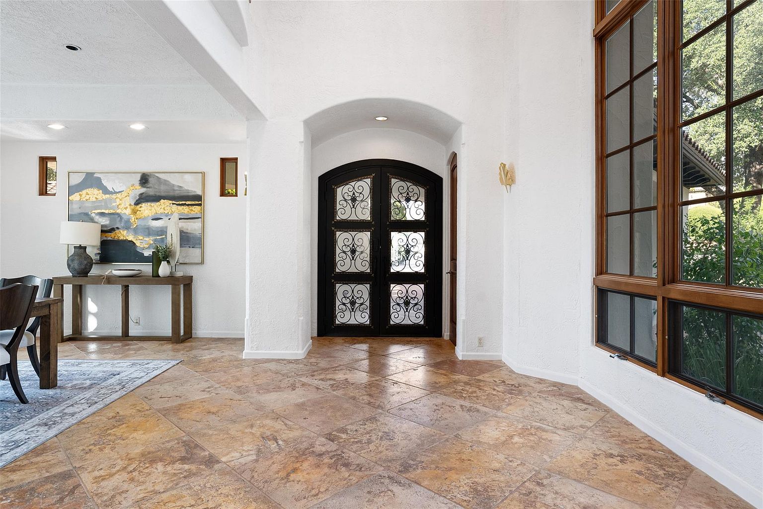 This interior shot showcases a grand entryway with travertine flooring and white textured walls. A decorative black wrought iron door is centered under an arched opening, while a large window with wood trim provides natural light. The space is open and inviting, suggesting a well-maintained and stylish home.