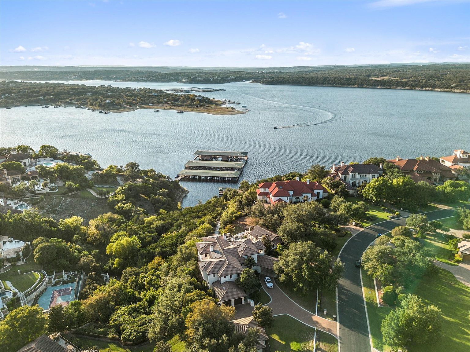 This aerial shot showcases a luxurious lakeside property with stunning views. The home features a tile roof, manicured landscaping, and a private driveway. The property also includes access to a boat dock on the lake, enhancing its appeal for water enthusiasts. The overall impression is one of exclusivity and high-end living.