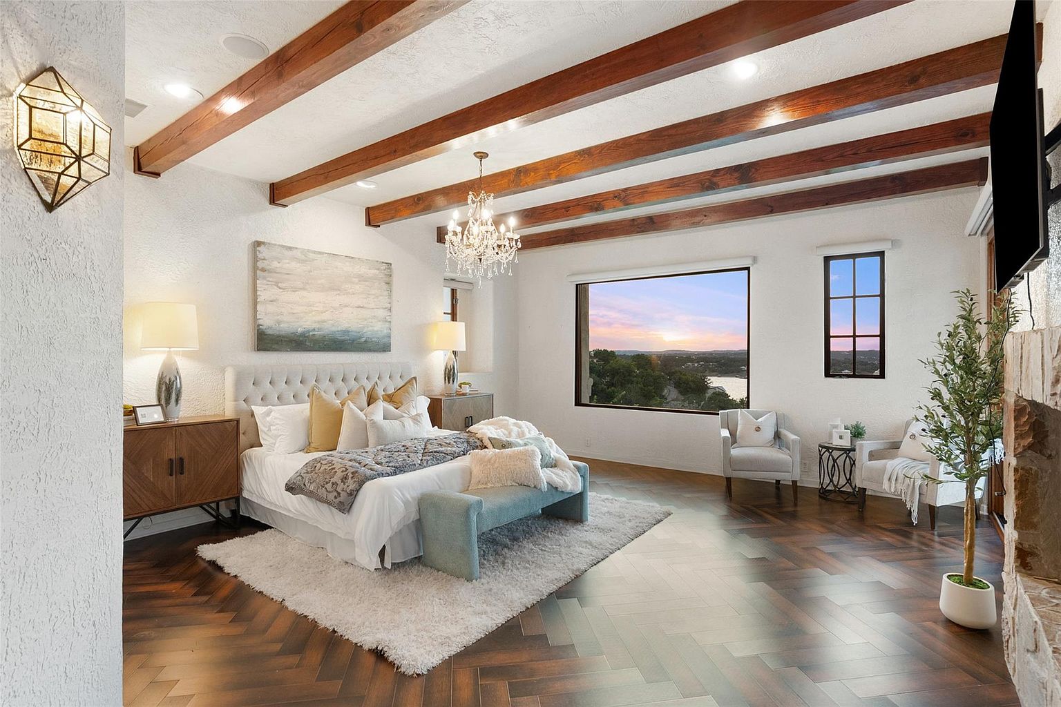 This is a luxurious primary bedroom featuring a king-size bed with a tufted headboard, complemented by wooden nightstands and elegant lamps. The room boasts exposed wooden beams on the ceiling, a chandelier, and a large window offering a scenic view. The herringbone wood flooring and plush rug add warmth and texture to the space, creating a sophisticated and inviting atmosphere.