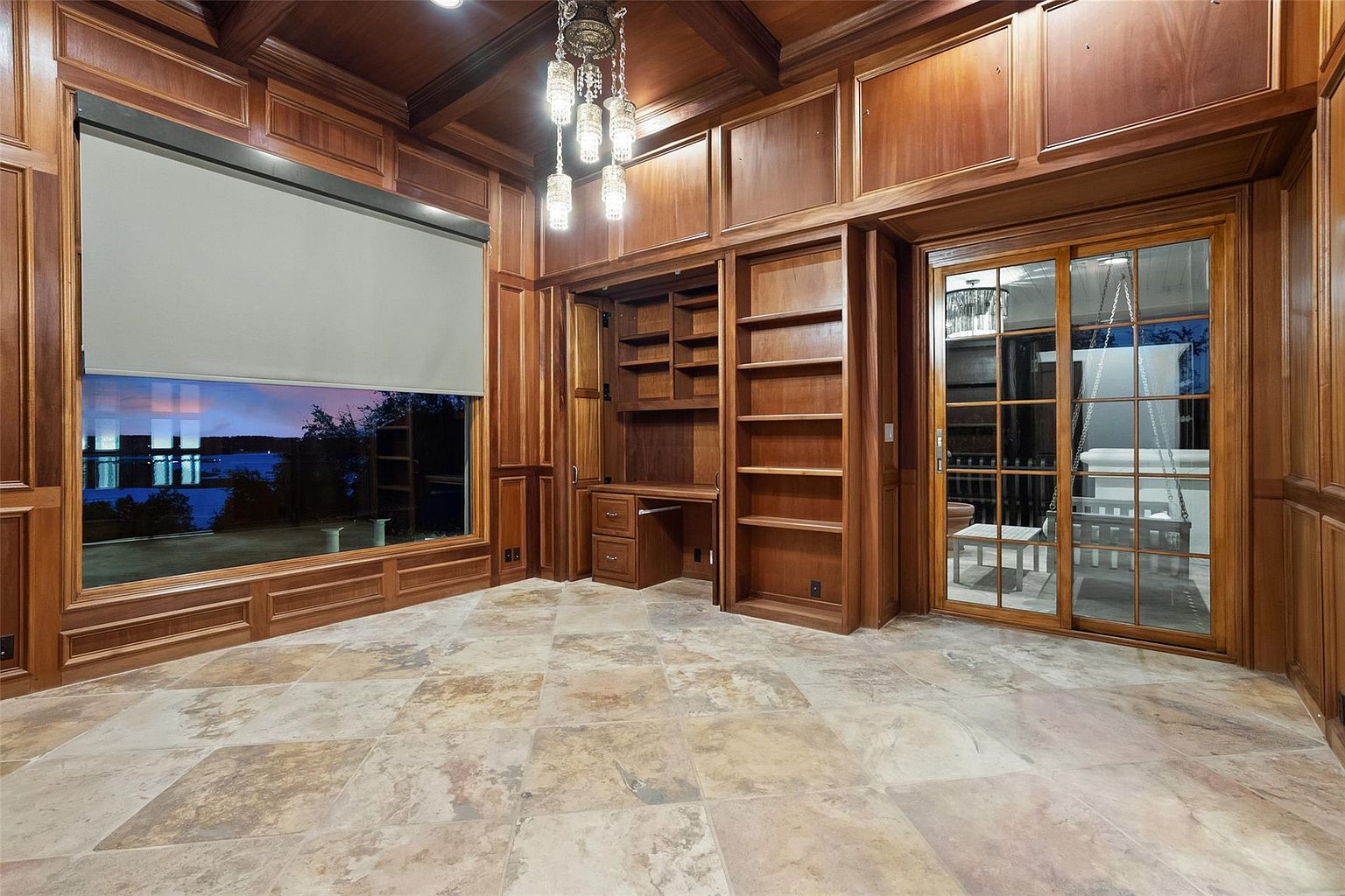 This is an interior shot of a luxurious home office or study, featuring rich wood paneling and built-in bookshelves. A large window offers a view of the water, while a chandelier adds a touch of elegance. The room is spacious with tile flooring, creating a sophisticated and comfortable workspace.