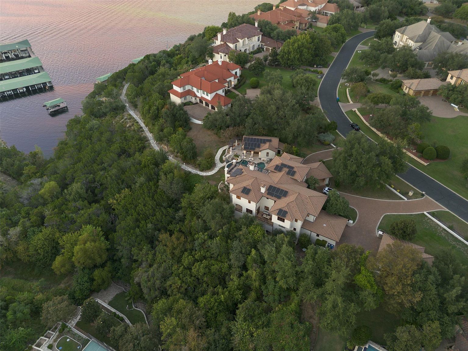 This aerial shot showcases a luxurious residential property nestled among lush greenery and overlooking a serene body of water. The house features a terracotta tile roof, multiple wings, and solar panels, suggesting a blend of classic style and modern sustainability. A winding stone path leads down to the waterfront, while a paved driveway connects the house to the street above.