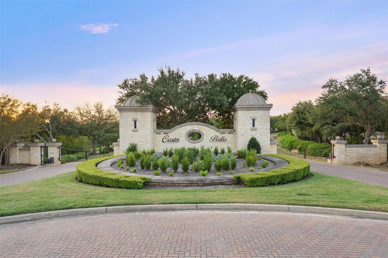 This image showcases the grand entryway to a community named "Costa Bella". The entrance features stone pillars topped with domes, a central stone sign, and a meticulously maintained circular garden with manicured hedges and various plants. The brick-paved road leads into the community, creating an inviting and upscale impression.