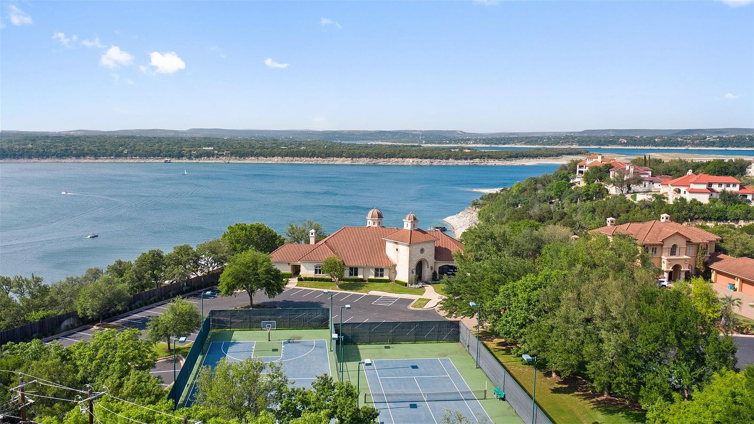 This aerial view showcases a luxurious estate with a terracotta-tiled roof, set against a backdrop of a serene lake and lush greenery. The property features well-maintained tennis and basketball courts, ample parking, and meticulously landscaped grounds. The overall impression is one of exclusivity and resort-style living.
